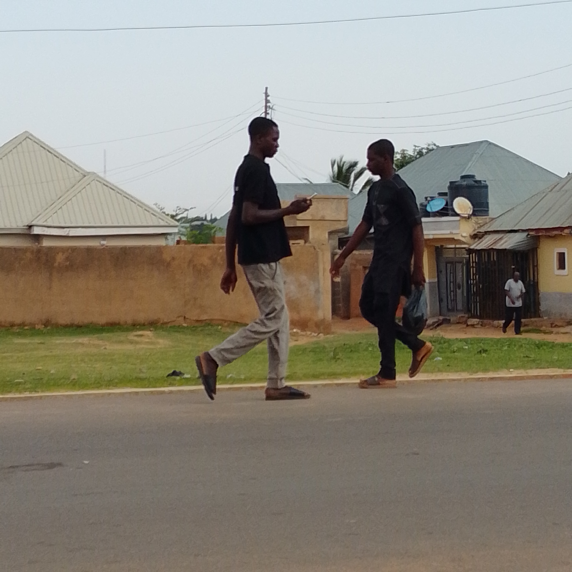 In Gombe, Nigeria, two young men walk along a paved road. The man on the left, wearing a black t-shirt and light grey trousers, holds a smartphone in his right hand and is looking down at it. The man on the right, dressed in a black traditional outfit, carries a dark bag in his left hand. Behind them, a low, brown wall borders a grassy area. Beyond the wall, a light-colored house with a corrugated metal roof stands, and further back, other buildings and palm trees are visible. Power lines are strung across the sky. A third man, wearing a light-colored shirt, is walking away from the buildings in the background. The sky is overcast.