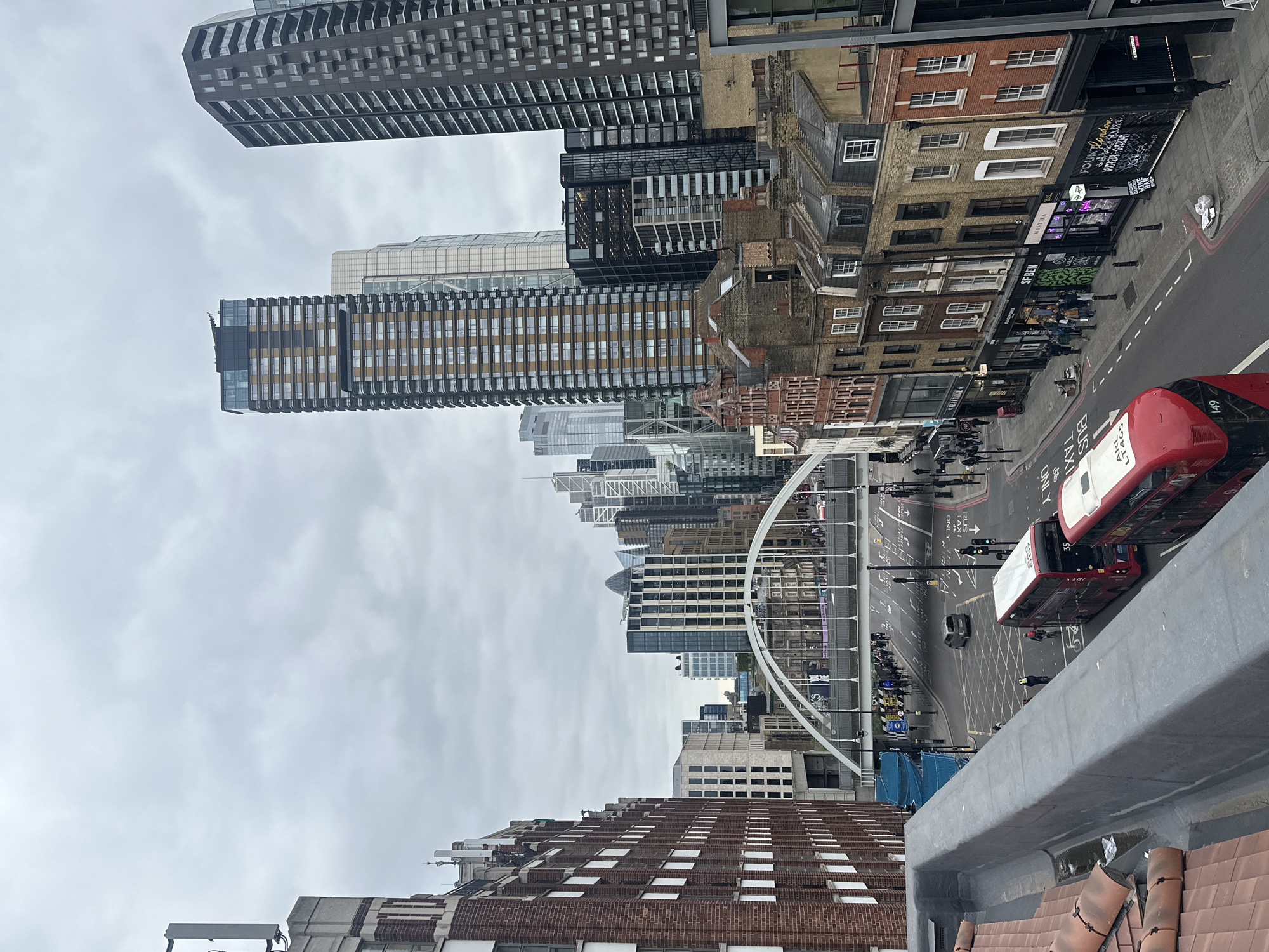 This is a ground-level, eye-level, medium shot of an urban cityscape, featuring buildings, streets, and pedestrian traffic. In the foreground is a section of roof with tiles. Beyond, streets are filled with cars, buses, and pedestrians. On the right side are two red double-decker buses, and other cars are also visible. The middle ground features a pedestrian bridge crossing a street. The bridge is a light color and spans multiple lanes. The background displays a mix of architectural styles, with modern skyscrapers juxtaposed against older brick buildings. The sky is overcast. The location appears to be a central area in London, United Kingdom.