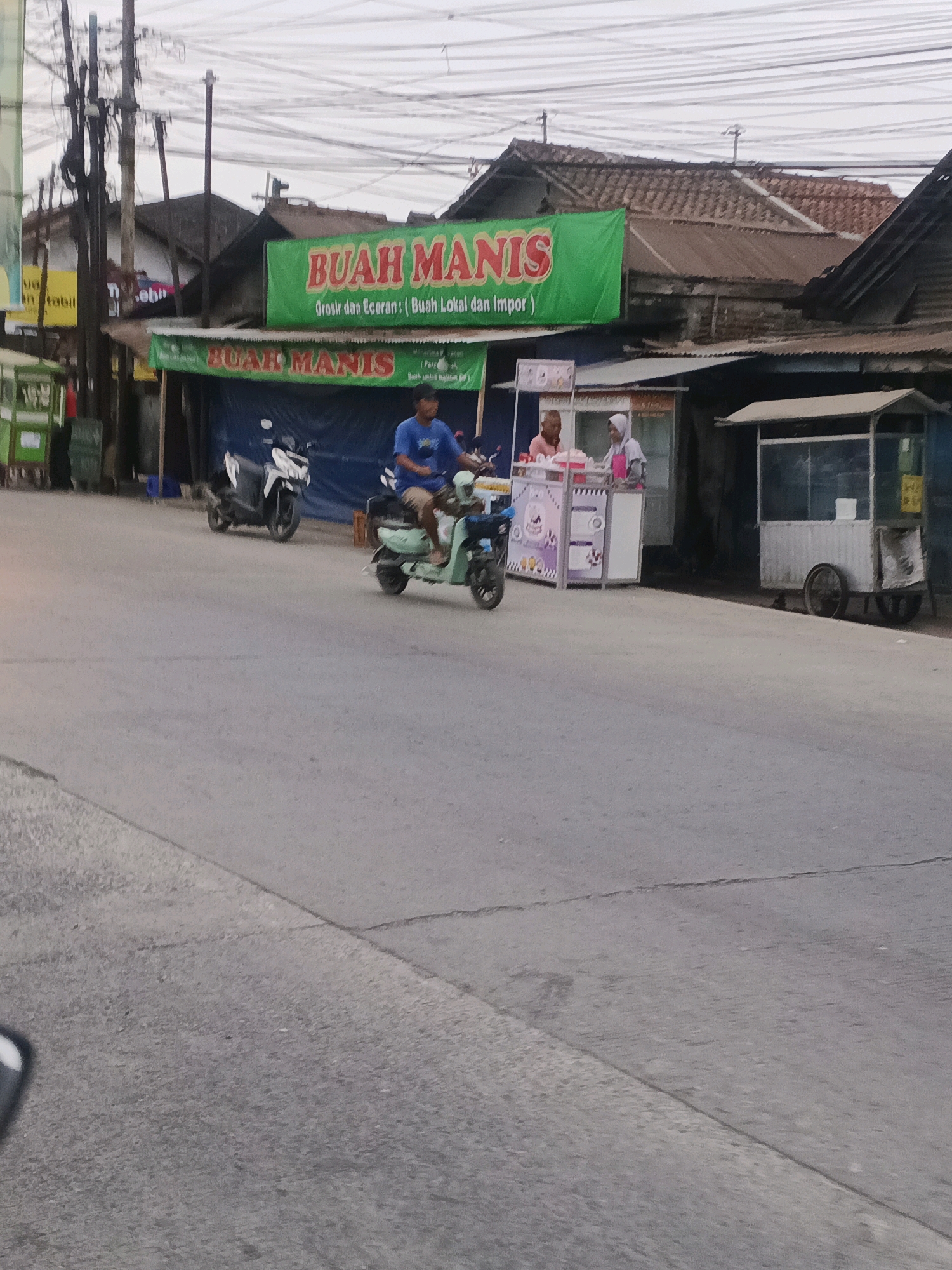 The image depicts a street scene in Kedung Menjangan, Indonesia. A man in a blue shirt and shorts is riding a light green scooter. In the background, there is a food stall with a pink and white striped counter, and two people are visible behind it. To the right, another stall with a white frame and a corrugated metal roof is visible. Several motorcycles are parked. Two green food carts are on the left side of the image. The road is paved and appears to be in moderate condition with some visible cracks. Overhead, a tangle of electrical wires spans the sky. A green banner with "BUAH MANIS" printed in red lettering is prominently displayed above the stalls.