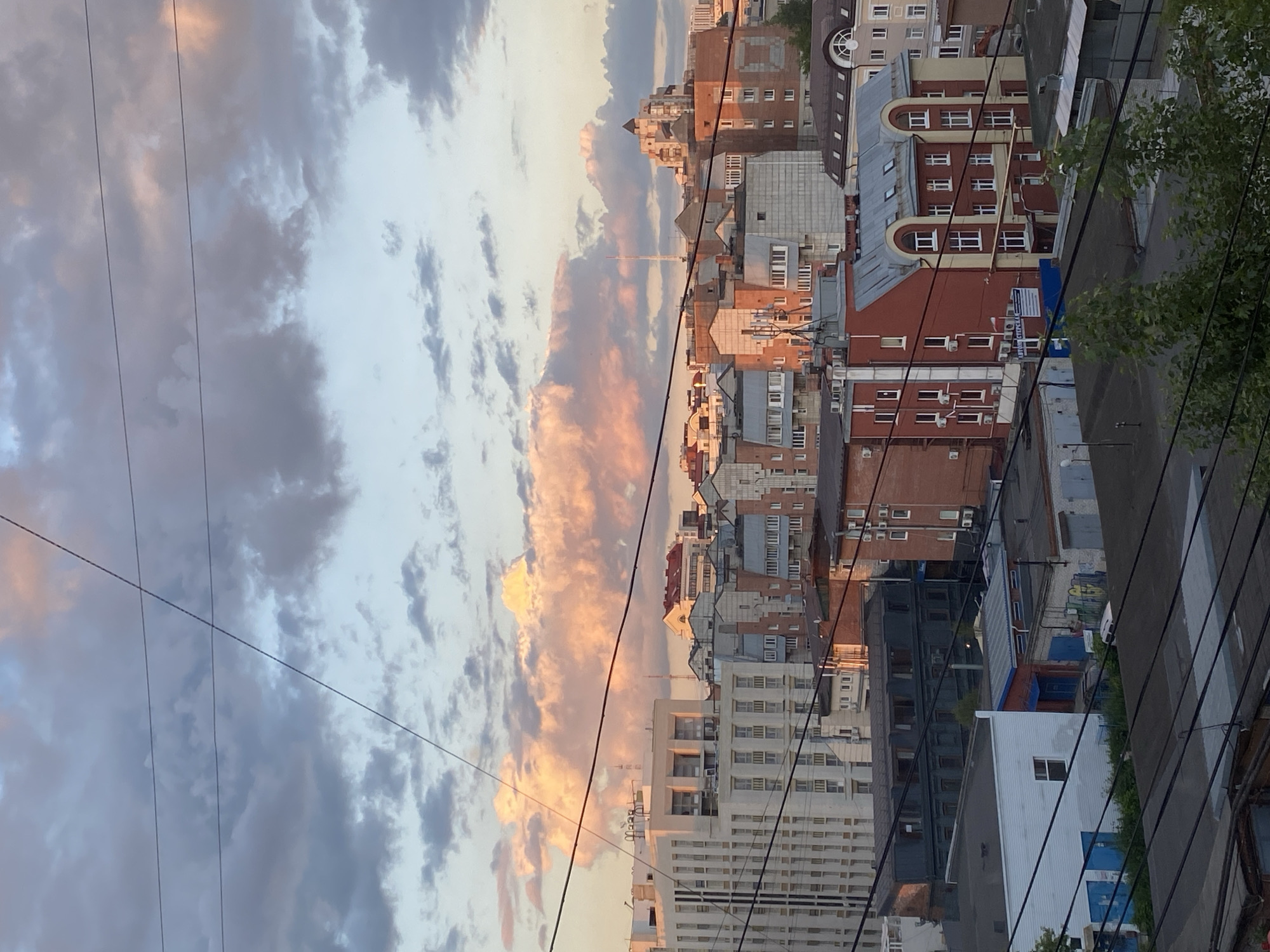 The image depicts an aerial view of a city skyline at sunset, with the sun casting warm hues on the clouds. The scene captures a mix of architectural styles, from modern high-rise buildings with light-colored facades to older brick structures with red and beige tones. Various rooftops are visible, some flat and industrial, others with pitched roofs. Power lines crisscross the frame, adding a sense of urban infrastructure. In the distance, a few taller buildings and spires punctuate the horizon. The overall impression is of a densely populated urban environment experiencing the end of the day.
While the image is a generic urban landscape, it is identified as being taken in the city of Perm, Russia. This information is provided contextually and not visually discernible from the photograph itself. The photograph does not contain any people, actions, or interactions. The subjects are primarily buildings and the natural elements of the sky and clouds. The environment is urban, characterized by dense construction and visible utility infrastructure.