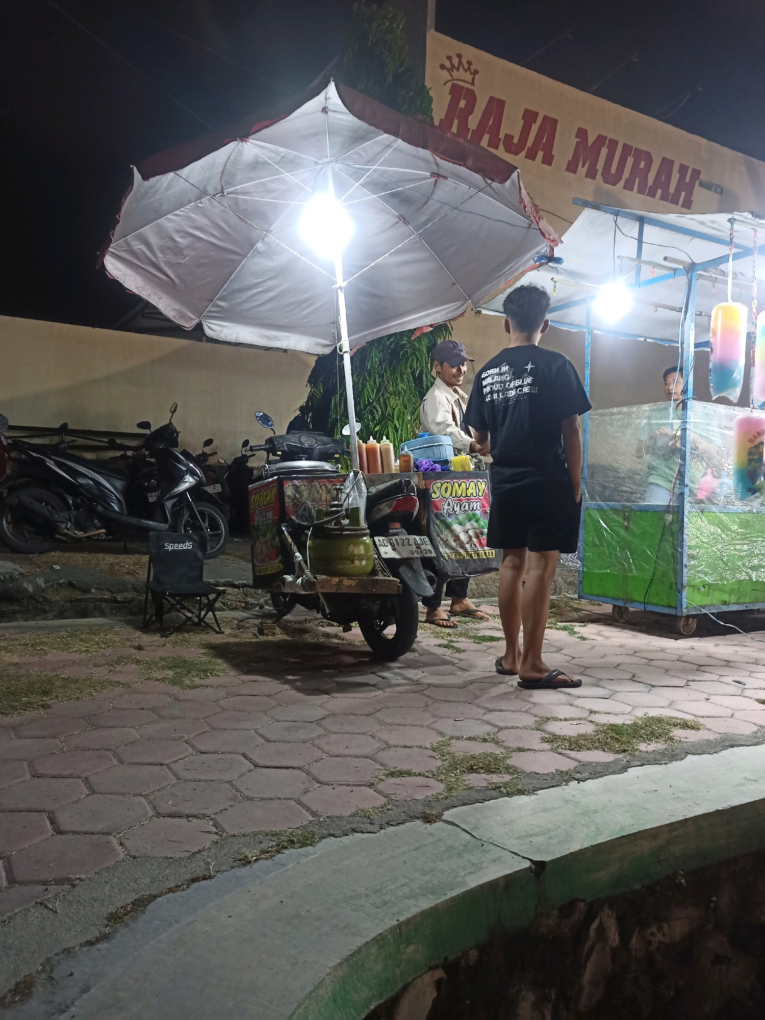 In Tanon, Indonesia, at night, a street food vendor operates under a large white umbrella. A young man wearing a black t-shirt and shorts stands facing away from the camera, his hands in his pockets. Next to him, a vendor serves customers from a cart selling "Somay Ayam". Several motorcycles are parked behind the vendor's cart. In the background, a sign reads "RAJA MURAH". The ground is paved with hexagonal tiles, and patches of grass are visible. The lighting appears to be from artificial sources.