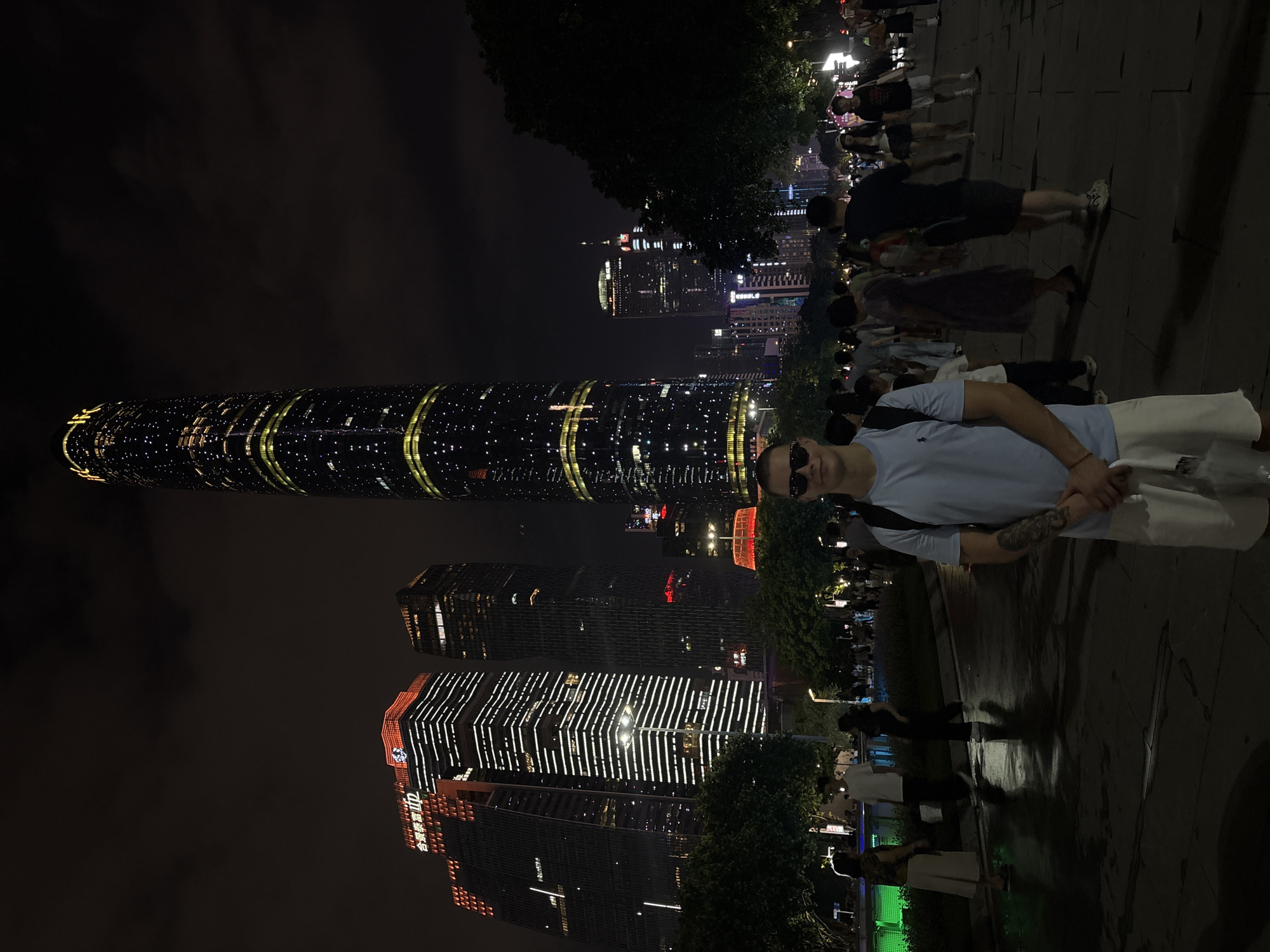 The image depicts a nighttime scene in Tianhe District, China, characterized by illuminated skyscrapers and a gathering of people. A young man, wearing sunglasses, a light blue t-shirt, and white shorts, stands in the foreground, facing the camera. He has tattoos on his left arm and is holding a white bag. Behind him, a multitude of people are walking and standing on a paved area, with trees and lush vegetation lining the path.
Dominating the background are several tall, modern skyscrapers, brightly lit with patterns of lights. One particularly slender skyscraper, possibly the Canton Tower, is distinguished by its cylindrical shape and illuminated facade, featuring horizontal bands of yellow and white lights interspersed with scattered white dots. Other buildings display various architectural styles and lighting designs, some featuring orange and white illuminations. The sky above is dark and cloudy, typical of a nighttime urban environment. The ground appears wet, reflecting the ambient light, suggesting recent rain or a water feature. The overall atmosphere suggests a bustling urban plaza or promenade in a metropolitan area.