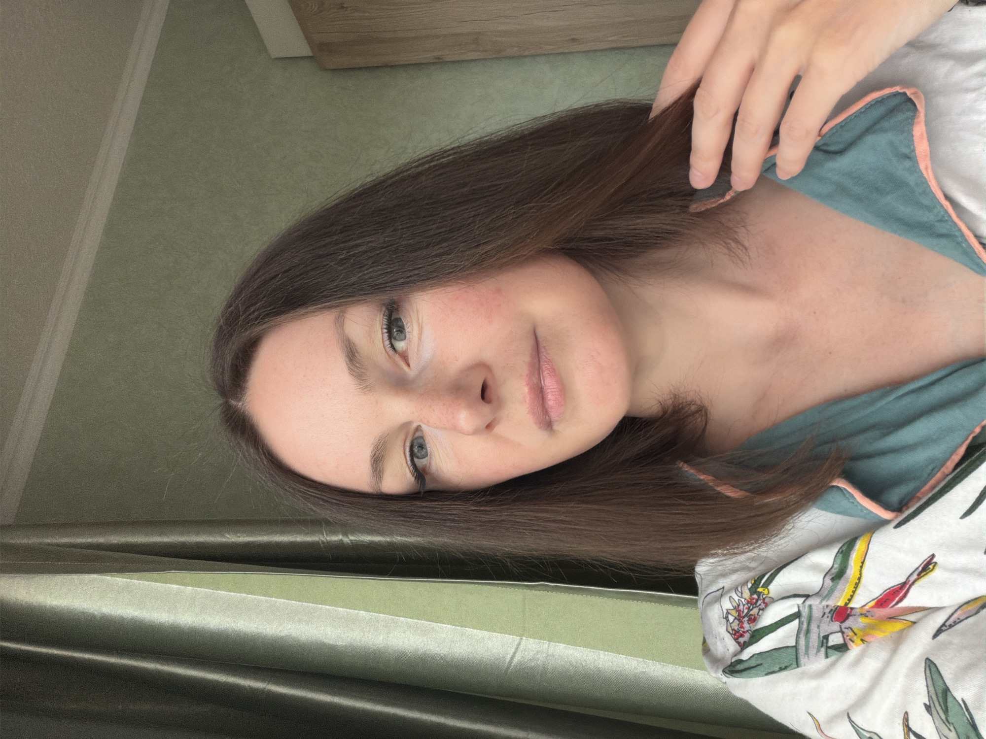 A woman with brown hair and light eyes poses for a selfie. She is wearing a patterned top with a green collar. The background includes a green wall and a wooden cabinet. The location is unknown.
