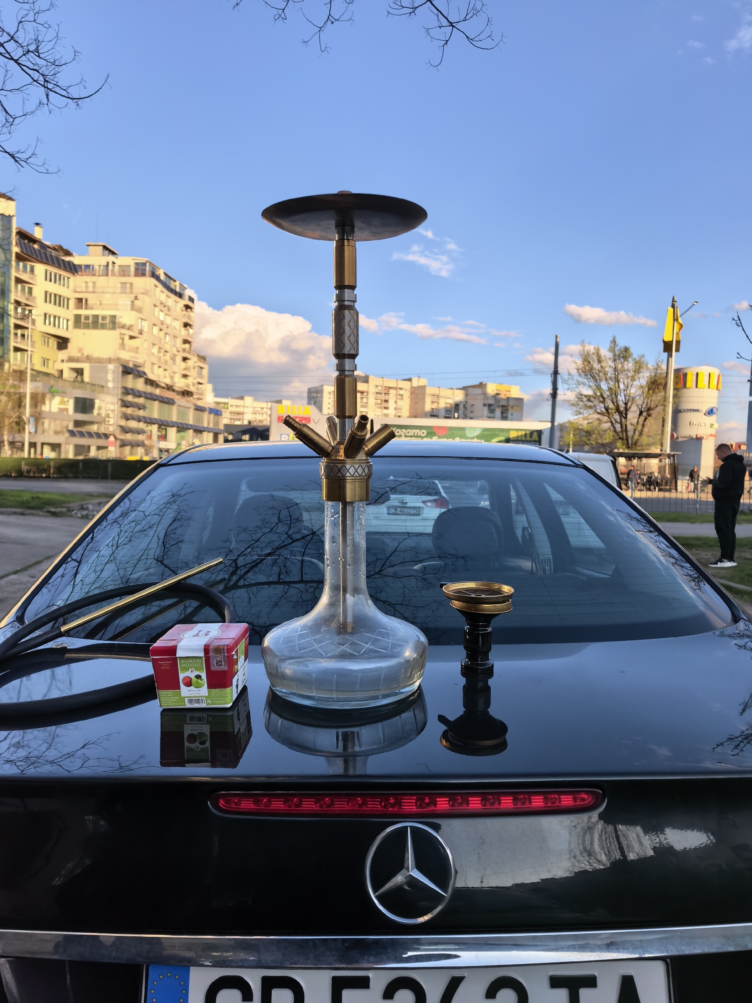 The image shows a hookah setup on the trunk of a black Mercedes-Benz car. The hookah is complete with a hose, bowl, and a pack of apple-flavored hookah tobacco. The car has a license plate from Bulgaria. In the background are buildings and a street scene that includes a pedestrian, trees, and signage. The location appears to be in Sofia, Bulgaria.