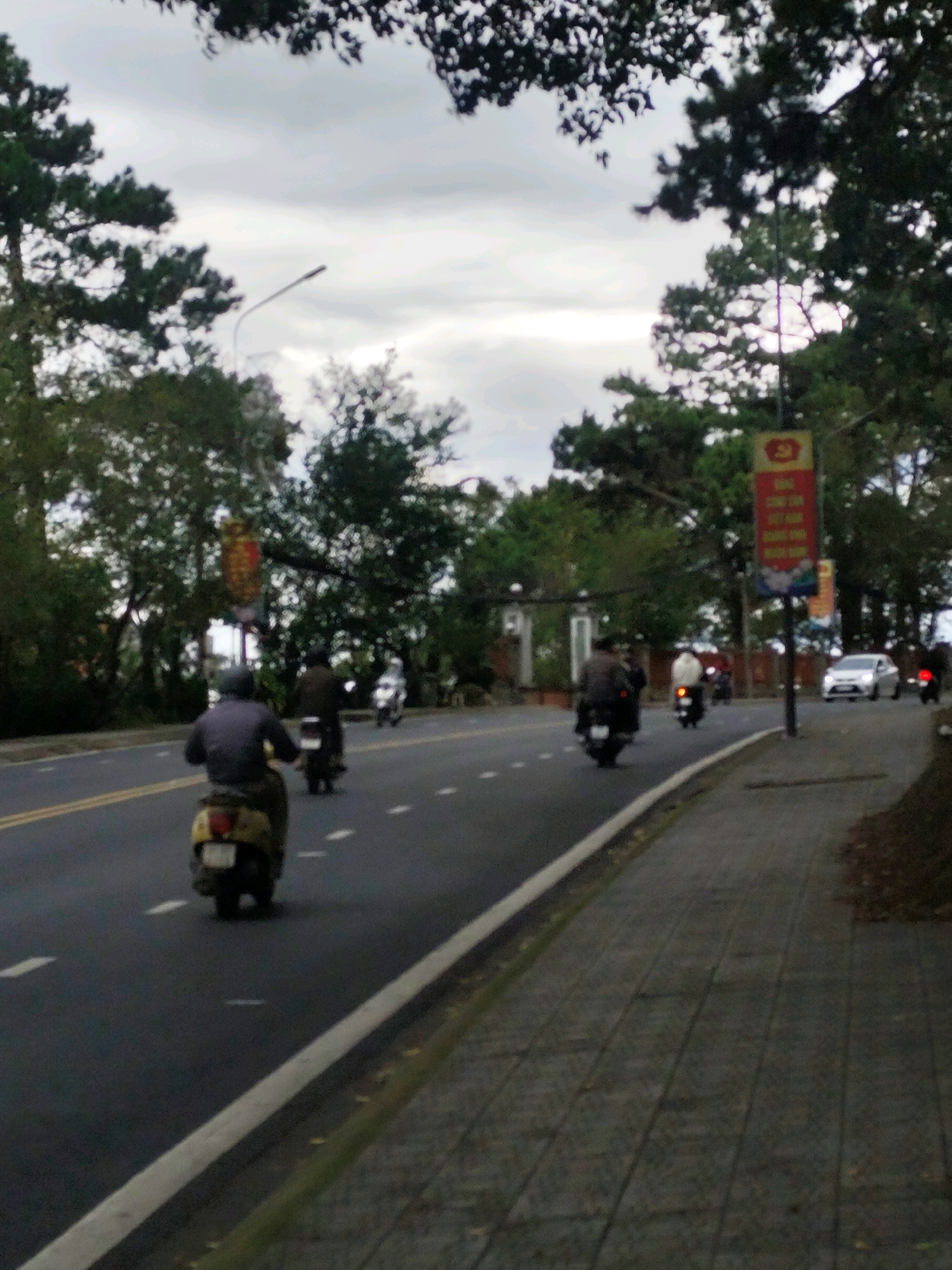 The image depicts a street scene in Da Lat, Vietnam, likely during daylight hours under an overcast sky. The road is paved, with yellow and white lane markings. Several people are riding motorcycles and scooters on the road, moving in the same direction. In the foreground, a person on a yellow scooter is visible from behind. Further down the road, other motorcycles and a white car are present. The roadside features a paved sidewalk, trees, and some foliage. A banner with red and yellow colors is hanging from a pole on the right side of the road. The overall impression is a typical urban or suburban street with active traffic.