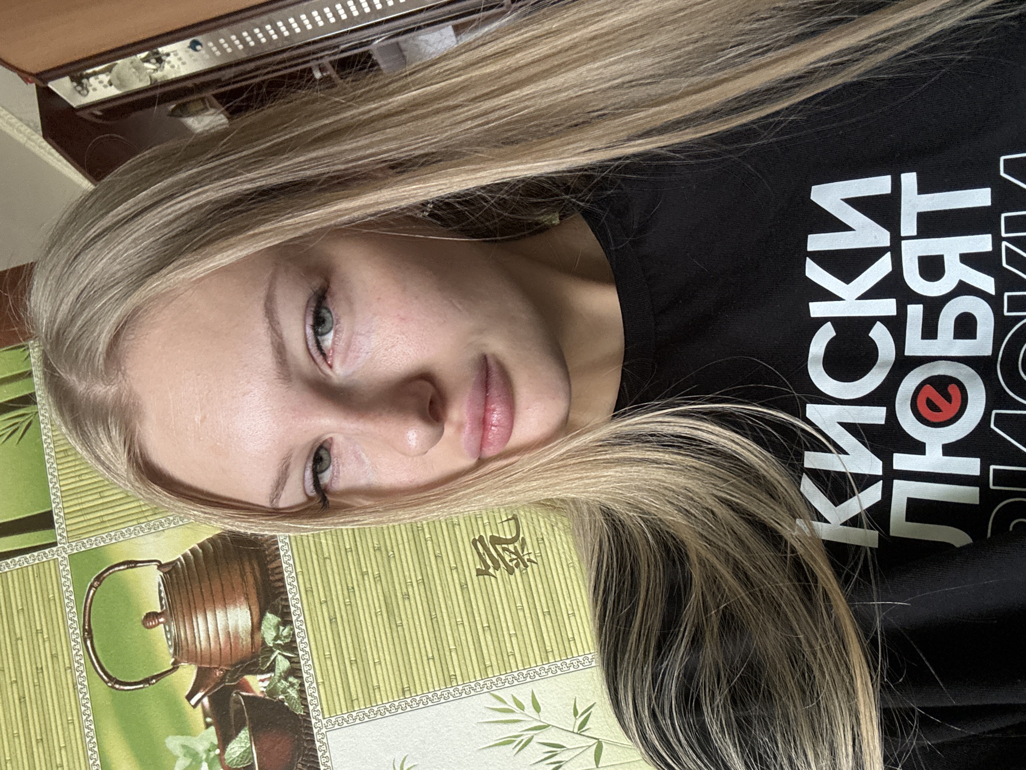 A young blonde woman with light blue eyes and fair skin is shown from the chest up, looking directly at the camera. She is wearing a black t-shirt with white Cyrillic lettering. The background appears to be a wall with a green bamboo-patterned wallpaper, and to the right, a wooden cabinet with visible shelves. The lighting is natural, coming from the front and slightly to the left, highlighting the woman's face and hair. There are no discernible signs or landmarks that would indicate the specific location within Комарихинский, Russia.