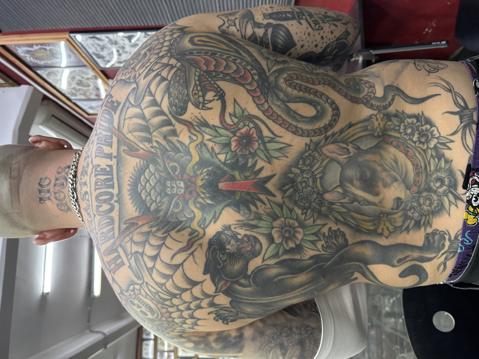 A man's back and shoulders are covered in numerous tattoos. The tattoos include a dragon, snake, dog, panther, skull, flowers and barbed wire. "HARDCORE PRIDE" is tattooed on the upper back. Another tattoo on the neck reads "NO GODS". The man has a chain around his neck. The tattoos cover the man's upper back, shoulders and upper arms. The scene appears to be inside a building, likely a tattoo studio. The location is not Moscow, Russia.