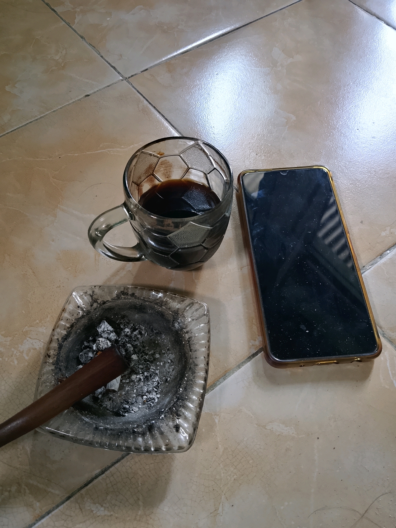 A high-angle medium shot shows a glass mug of dark liquid, an ashtray with cigarette butts and ashes, and a smartphone with a gold-colored frame placed on a beige tiled floor. The mug is partially filled with a dark liquid, possibly coffee, and has a textured pattern. The ashtray is square with rounded corners and contains several cigarette butts and ashes. A dark brown, unlit cigar lies horizontally across the ashtray. The smartphone is dark and appears to be off, with visible dust on its screen. The tiled floor exhibits a subtle cracked pattern. The scene is set indoors, likely in Jember, Indonesia, given the context. There are no people or explicit interactions depicted. The lighting suggests an indoor environment with ambient light.