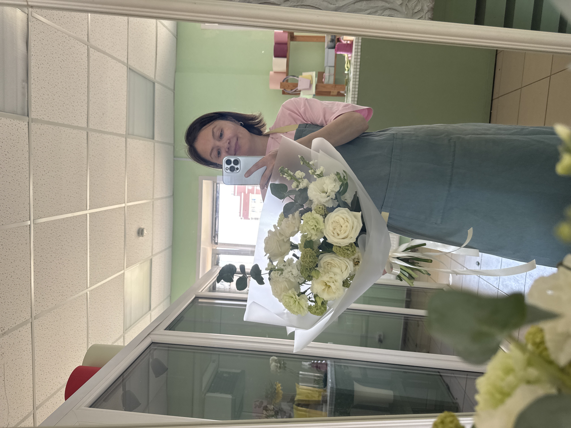 A woman is reflected in a mirror, holding a bouquet of white flowers and taking a selfie with a silver iPhone. The woman has short brown hair and is wearing a pink shirt under a blue-green apron. The bouquet is wrapped in white paper. The reflection shows a room with mint green walls and a grid ceiling. In the background, shelves display rolls of colored paper. Through a window visible in the mirror's reflection are buildings, suggesting a cityscape. The city is Izhevsk, Russia.