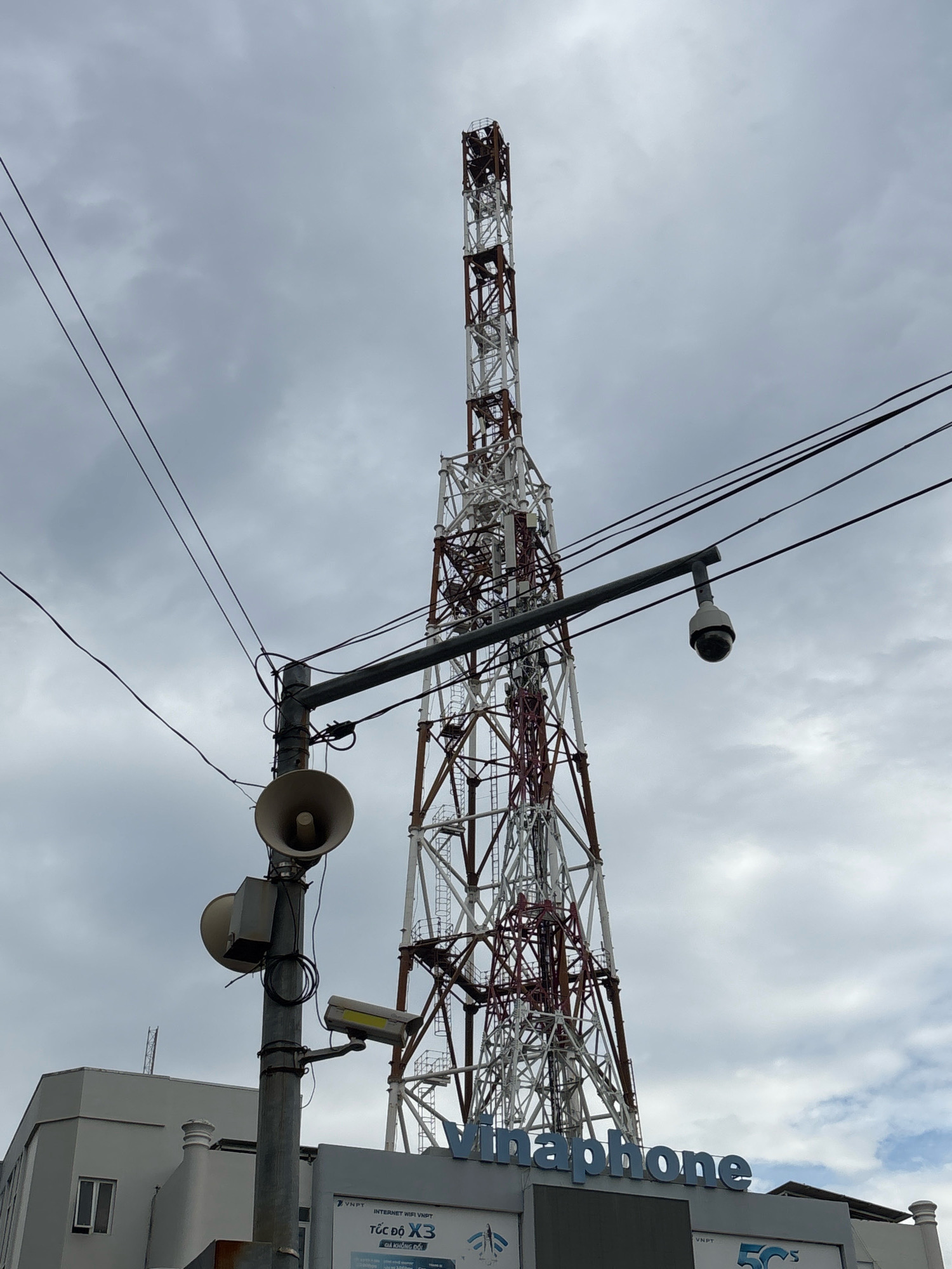 An outdoor, eye-level, medium shot captures a towering cell phone mast against a cloudy sky in Nha Trang, Vietnam. The mast, constructed of white and red lattice steel, dominates the frame. Attached to a pole in the foreground are two circular, bell-shaped speakers, one above the other, and a security camera. Another security camera is mounted on an arm extending from a horizontal bar above the speakers, positioned closer to the mast. Wires crisscross the sky in front of the mast. A low-rise building with a light grey facade is visible to the left of the mast. The building features a rectangular window and a white decorative element on its roof. A sign with blue lettering, "Vinaphone," is visible on the building's facade. The overall impression is one of an urban infrastructure scene.