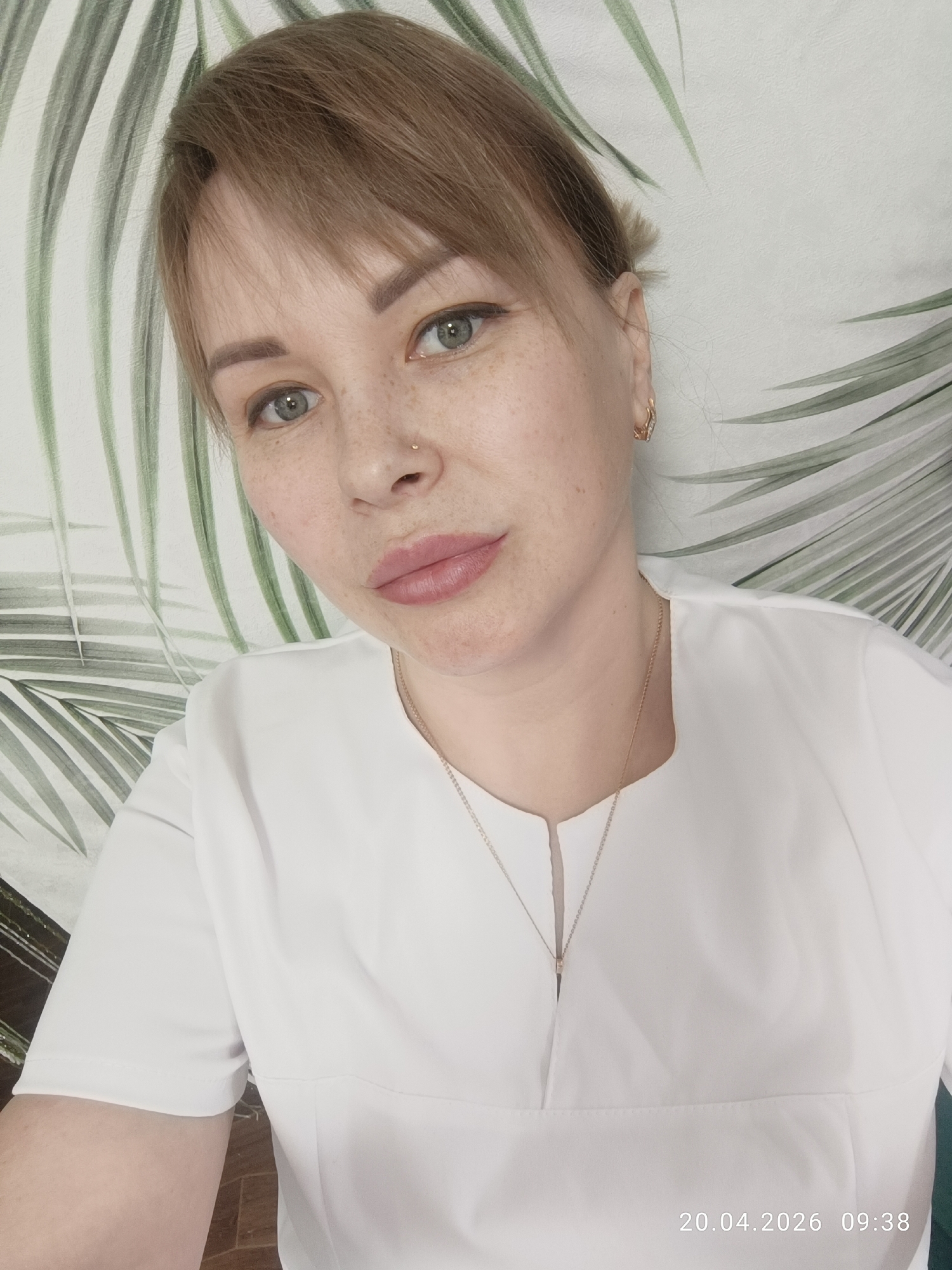 A person with light skin, freckles, and short brown hair is facing the camera, wearing a white top and a gold necklace. They have a nose piercing and gold earrings. The background features a wallpaper with a design of green palm leaves. The date "20.04.2026 09:38" is visible in the bottom right corner. The location is Chaikovsky, Russia.