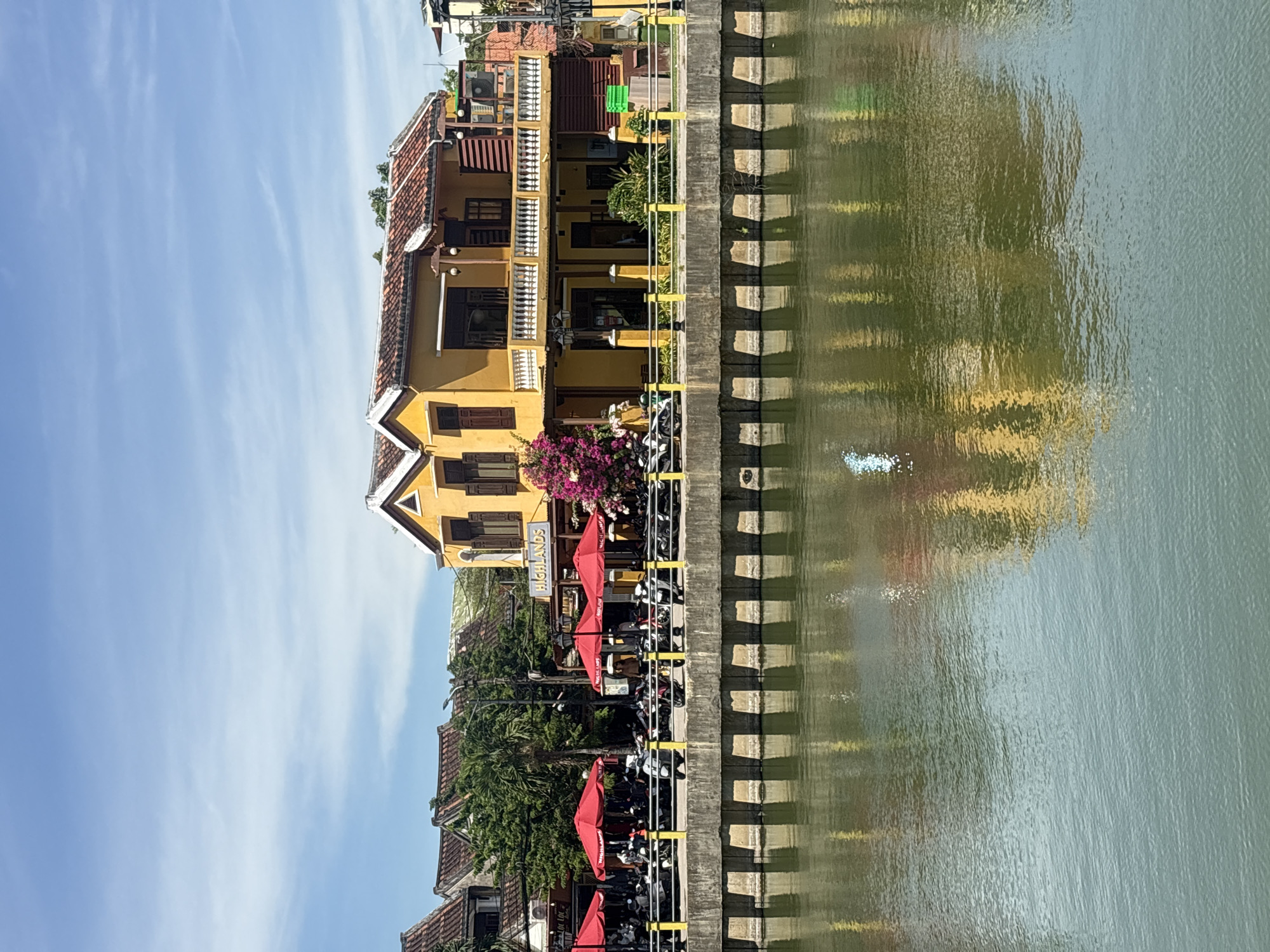 The image shows a yellow building with a red tiled roof in Hội An, Vietnam, located on the bank of a river. The building has multiple windows and a balcony with white railings. In front of the building, there is a row of red umbrellas covering outdoor seating areas. Many motorcycles are parked along the waterfront. The river in the foreground reflects the building and the sky. The sky is blue with scattered white clouds. The overall scene depicts a bright, sunny day in a bustling riverside area.