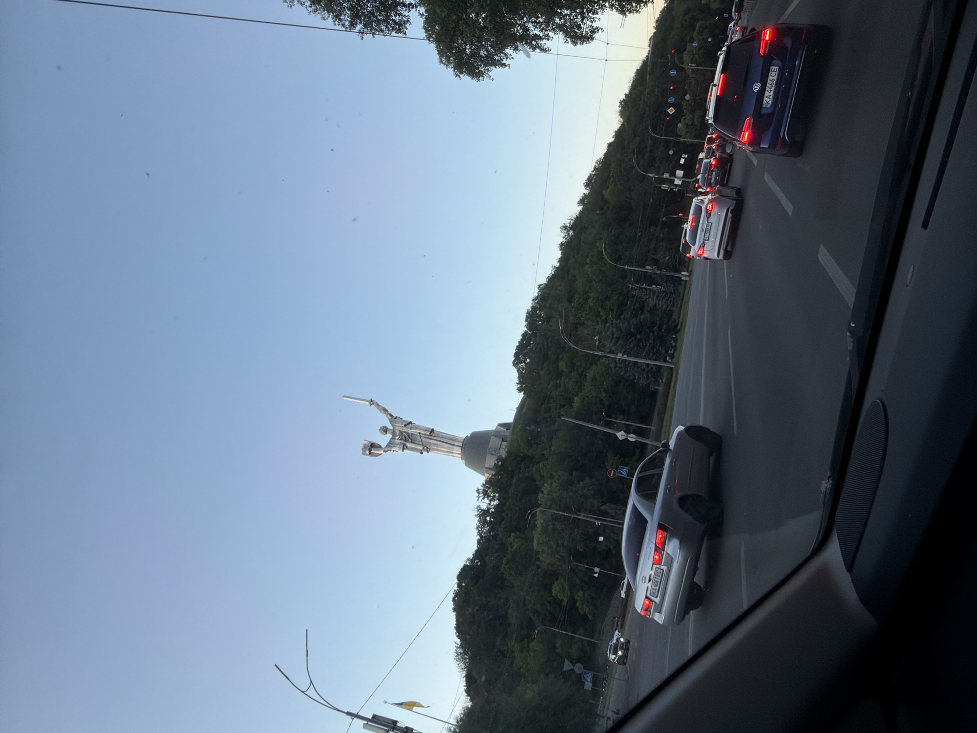 The image shows a wide-angle view of a street scene in Kyiv, Ukraine, from the perspective of a vehicle's interior. The main focal point is the Motherland Monument, a colossal statue of a woman holding a sword and a shield, prominently situated on a verdant hill overlooking the city. The monument is made of stainless steel and is a significant landmark of Kyiv.
The street is busy with traffic, with several cars visible. In the foreground, a silver sedan is turning, while other vehicles, including a blue Volkswagen, are driving in the same direction. Traffic lights and road signs are visible, indicating a well-developed urban infrastructure. The sky is clear and blue, suggesting fair weather.
The scene appears to be captured during daylight hours, possibly late afternoon or early evening, given the ambient light conditions. The overall impression is of a dynamic urban environment with a monumental historical site serving as a backdrop to everyday life.