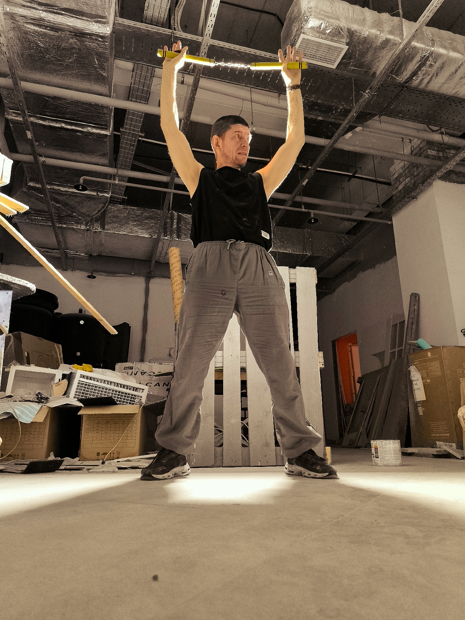A man with short, dark hair and a goatee stands in a construction or storage area, holding a yellow illuminated bar above his head with both hands. He wears a black t-shirt and grey sweatpants. The environment is characterized by exposed ceiling ductwork, pipes, and electrical conduits. Debris, including cardboard boxes, wooden planks, and other construction materials, is scattered on the concrete floor. The lighting suggests artificial sources, casting shadows on the floor and walls. The scene appears to be an unfinished indoor space.