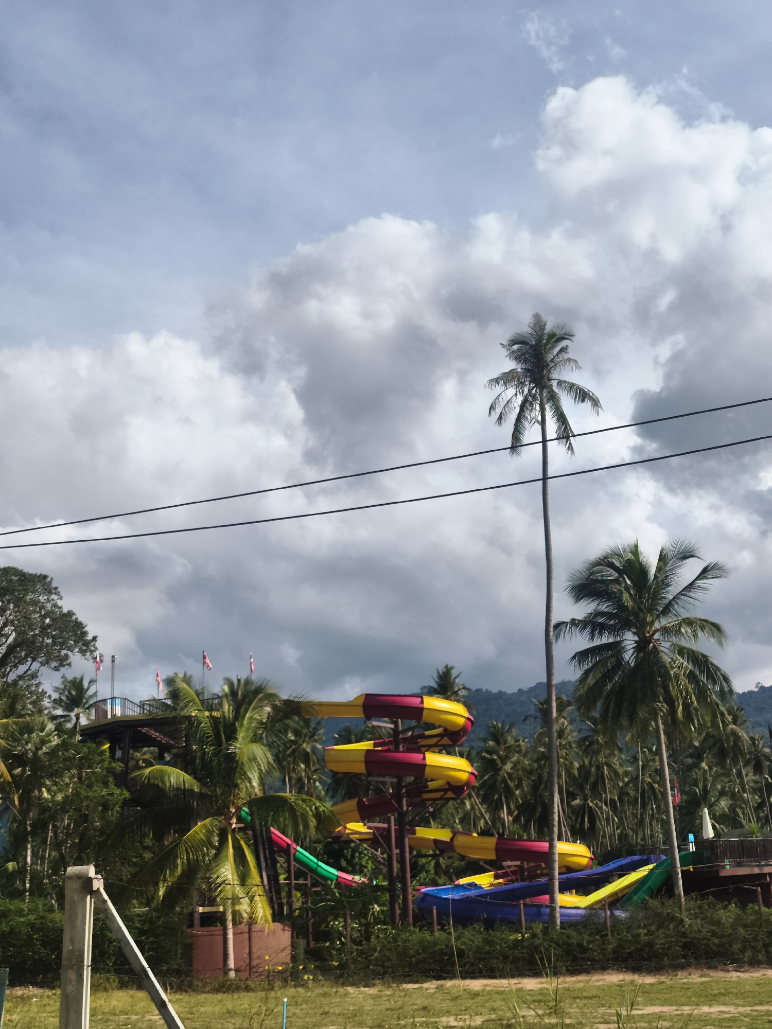 This is a vertical, medium-distance, eye-level shot of a water slide in a tropical setting. The water slide structure, comprised of colorful tubular sections in yellow, red, pink, green, and blue, is the primary focus. It is surrounded by lush green palm trees and dense foliage. In the background, a hazy mountain range is visible beneath a dramatic sky filled with dark, swirling clouds. Power lines stretch across the upper portion of the frame, with a tall, slender palm tree dominating the mid-ground to the right. A structure with flags is visible on the left, and a portion of a concrete pillar and fencing is in the foreground on the left. The scene appears to be outdoors in a humid climate, possibly a water park.