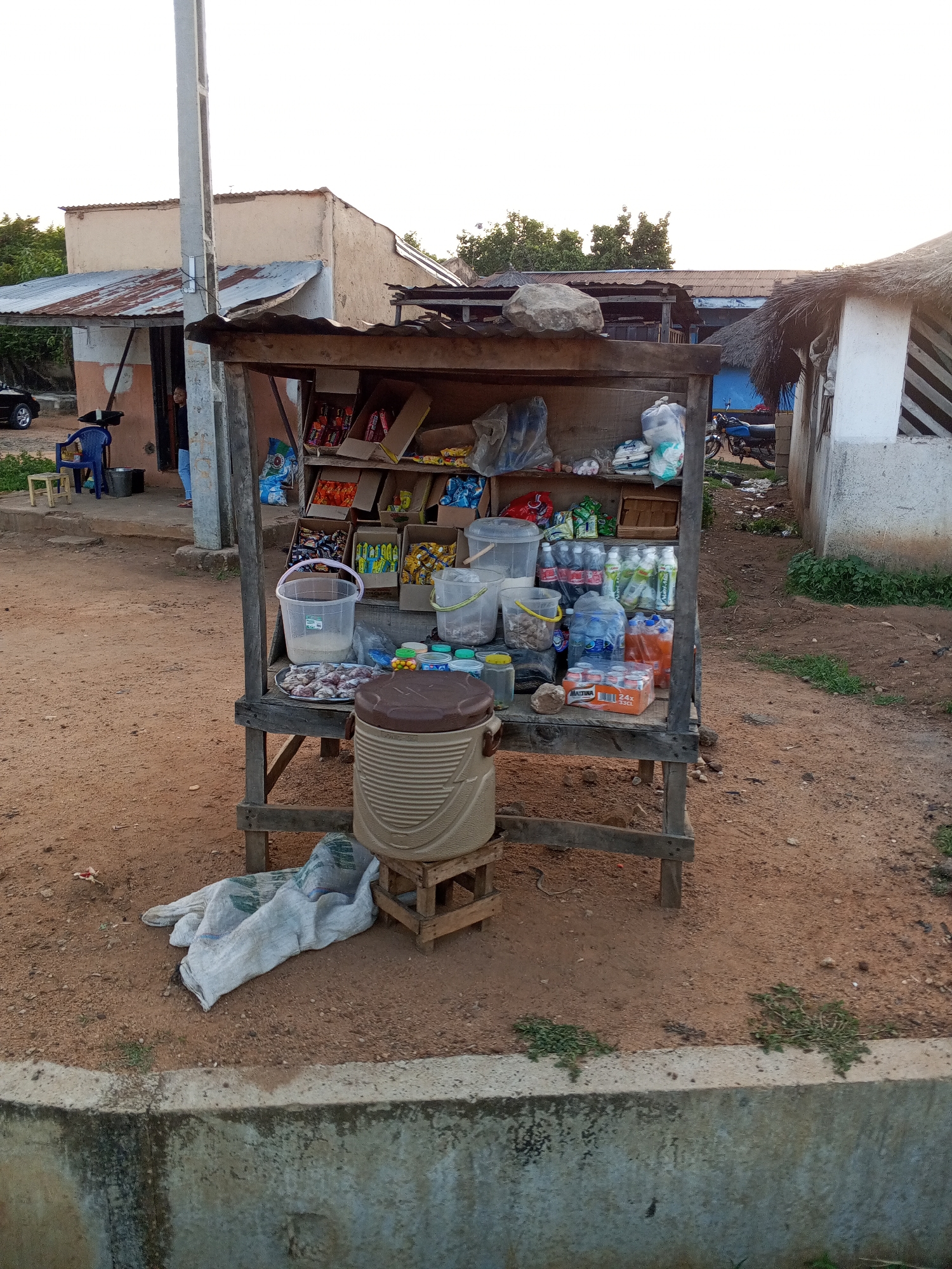 A street-side vendor stall is located in an unidentifiable city. The stall, constructed of wood and topped with corrugated metal, displays an assortment of goods, including snacks, drinks, and packaged foods. A large insulated water container rests on a small wooden stool in front of the stall. A white cloth bag is lying on the ground nearby. The ground is dirt. In the background, various buildings and a motorcycle are visible. A utility pole stands next to one of the buildings.
