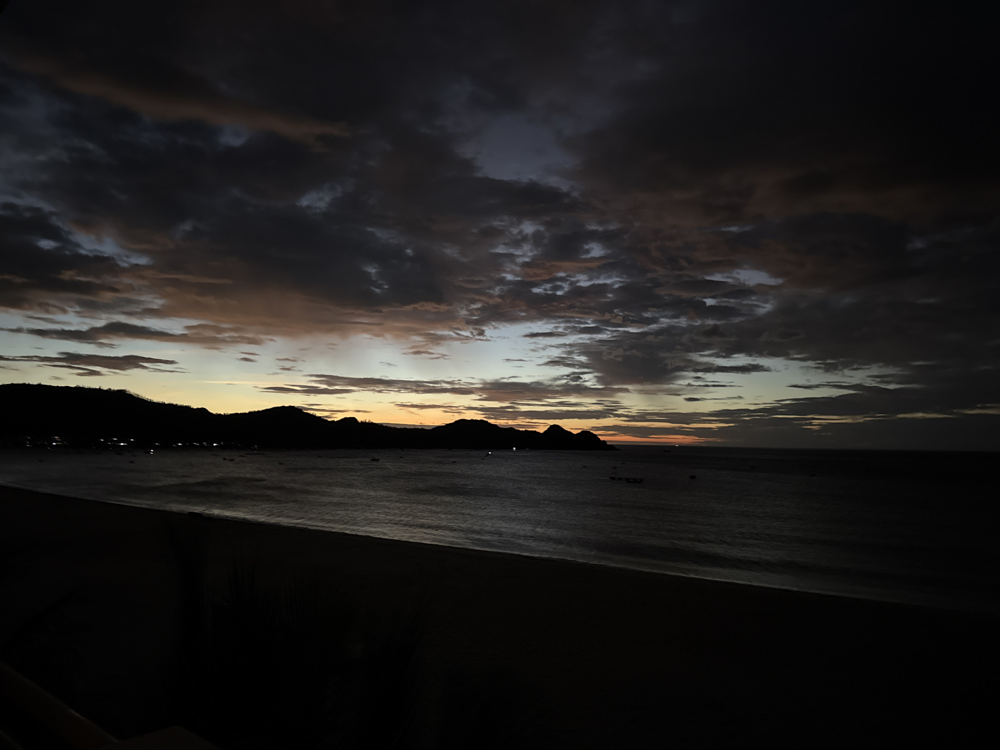 This image depicts a nighttime coastal scene in Song Cau, Vietnam, likely captured from an elevated vantage point. The foreground is dominated by a dark, silhouetted expanse, possibly a balcony or a rooftop, with minimal discernible features. Beyond this, a dark beach stretches out, meeting the calm, dark waters of the ocean. In the distance, a line of silhouetted hills or mountains forms the horizon, with faint lights suggesting a town or settlement nestled along the coastline. The sea is dotted with a few small, dark shapes that could be boats. The sky above is a dramatic display of twilight, with dark, heavy clouds interspersed with streaks of orange and pale blue, indicating the lingering light of sunset or the approach of dawn. The overall mood is serene and somber, characteristic of the transition between day and night.