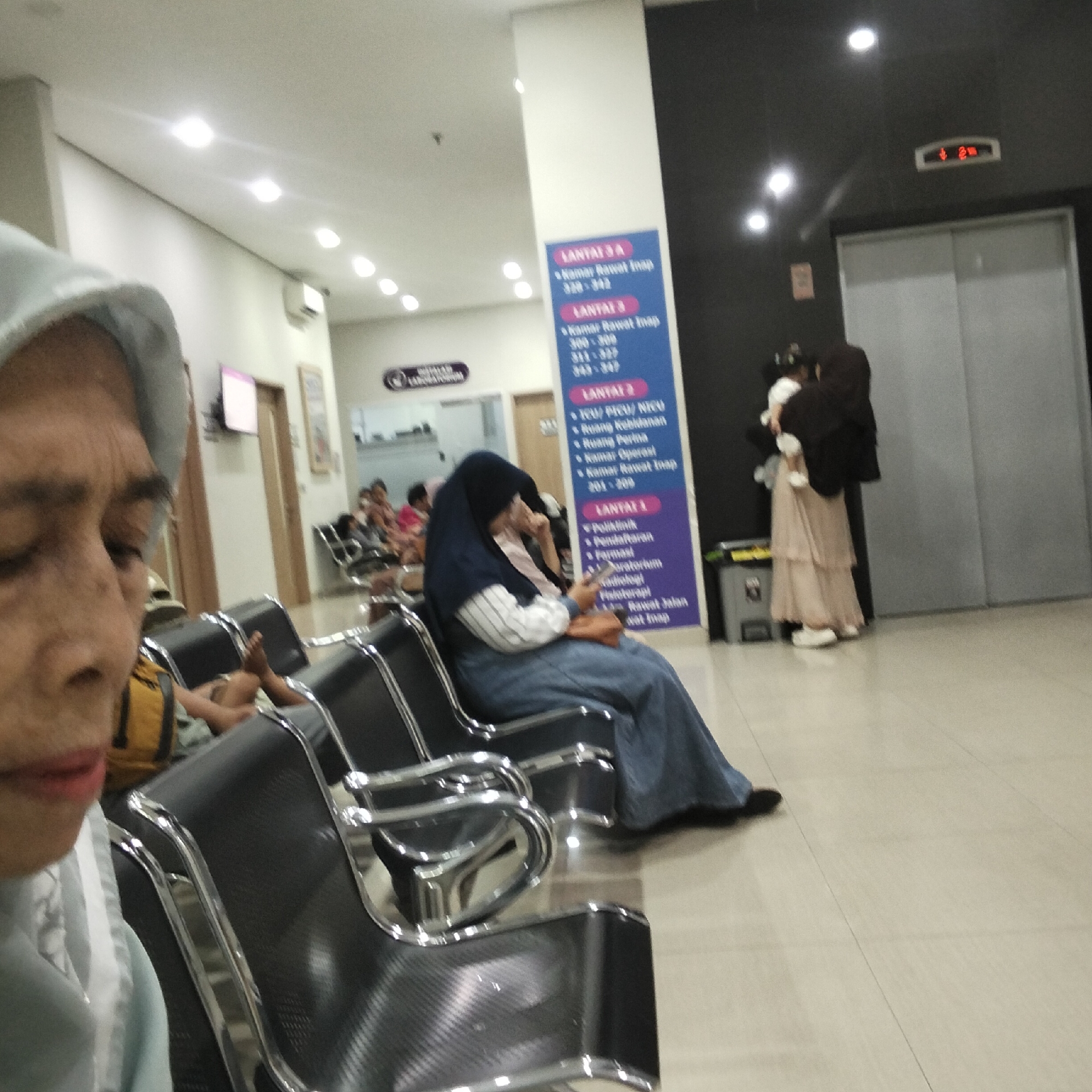 The image depicts a waiting area, likely in a hospital or clinic, with several people seated on dark, modern chairs. In the foreground, a woman's face is visible, with her head covered by a light blue hijab. She appears to be looking down. Further back, other individuals are seated, some obscured by the foreground subject. A blue sign with white text lists floor levels and room numbers, indicating "LANTAL 3 A", "LANTAL 3", and "LANTAL 2". The environment appears clean and well-lit, with a tiled floor. In the background, there's an elevator with a digital display showing "4" and "2". To the right of the elevator, a person in a dark outfit is holding a baby, while another person in a light-colored, tiered skirt stands beside them. The overall impression is that of a public waiting space within a medical facility in Sukabumi, Indonesia.
