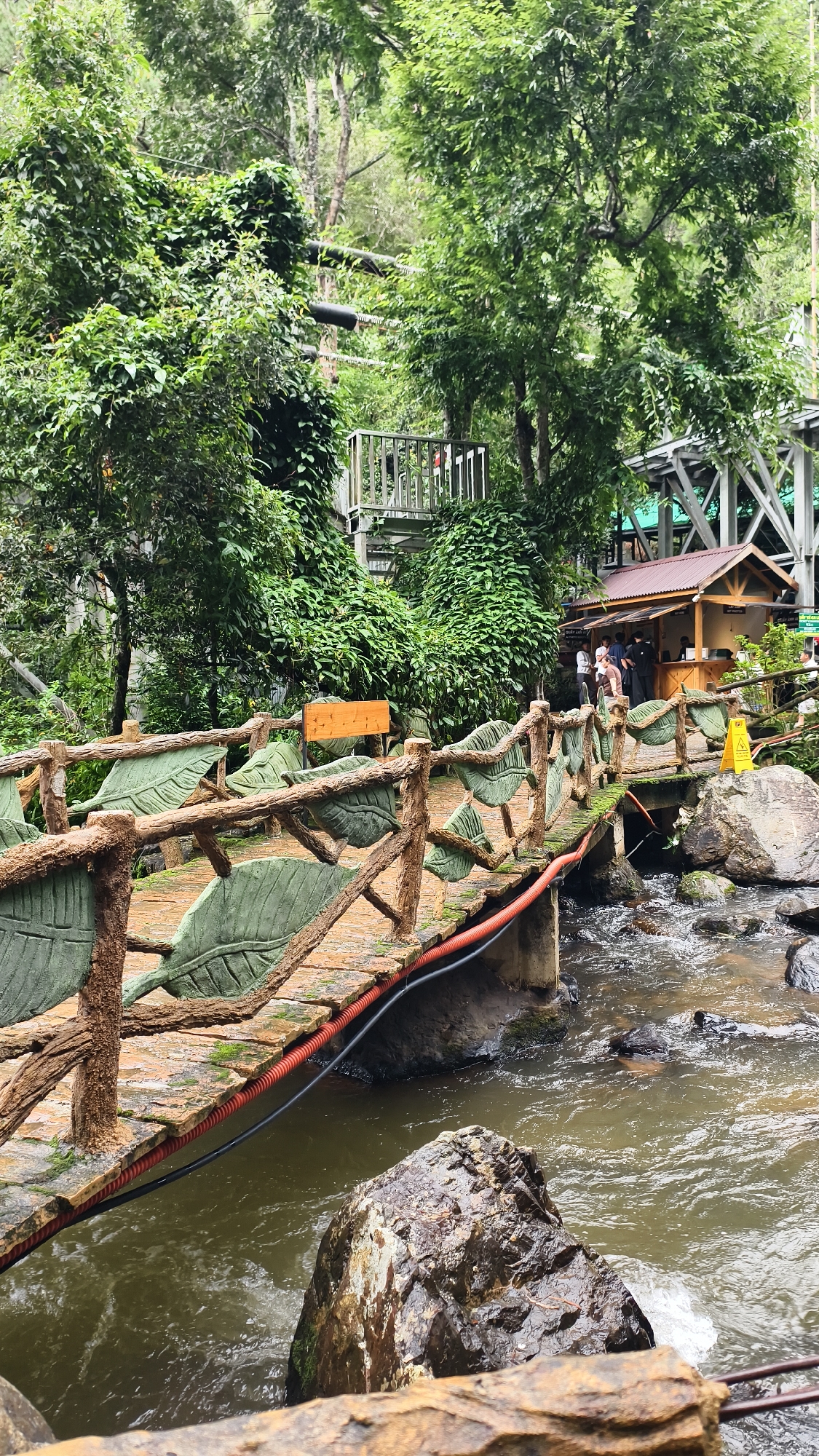 The image captures a picturesque scene in Da Lat, Vietnam, specifically at the Datanla Waterfall, a popular tourist destination known for its natural beauty and adventure activities. In the foreground, a decorative bridge with a rustic, wooden railing adorned with large, green, leaf-like carvings spans over a flowing stream. The bridge has red and black cables running along its side. The stream below is rocky and turbulent in parts, with clear water rushing over stones. In the background, lush green foliage and trees dominate the landscape, indicating a natural, forest-like environment. A small wooden hut, resembling a food stall or shop, is visible on the right side of the bridge, with several people standing near it. There is a yellow warning sign placed near the hut. Further in the background, there are glimpses of elevated structures, possibly part of a ziplining or adventure park setup, consistent with the activities offered at Datanla Waterfall. The overall atmosphere is one of a vibrant natural park with tourist amenities.