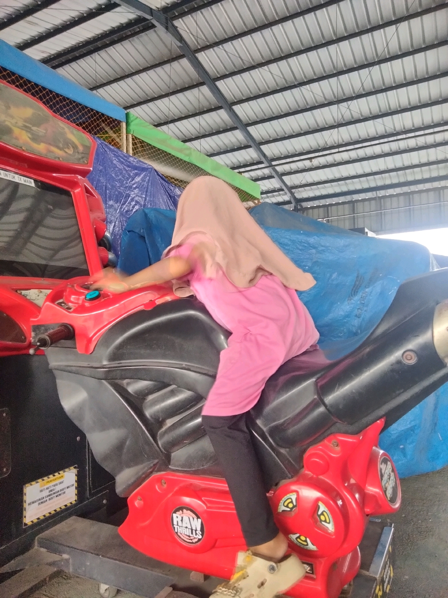 A person wearing a pink shirt and a tan hijab sits on a red and black toy motorcycle. The motorcycle is part of an arcade game with a screen and controls. The game appears to be inside a covered area with a metal roof supported by beams. Blue and green tarps are visible in the background, along with a net enclosure. A yellow and black caution sticker is affixed to the side of the game. The floor is concrete. This scene is likely located at an amusement park or arcade in the city of Sukaasih, Indonesia.