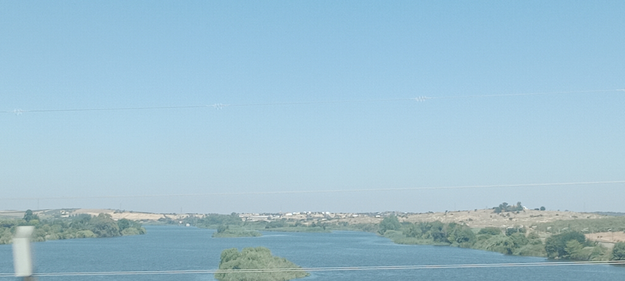 This is a wide-angle, eye-level, daylight, outdoor shot of a wide river, possibly the Guadiana River, as seen from a bridge or elevated position. The river occupies the lower half of the frame and is a deep blue color. The banks of the river are lined with lush green trees and vegetation, with some small islands of greenery visible within the water.
In the distance, a town or city is visible on the rolling hills beyond the river. The buildings are small and scattered, with some appearing to be white or light-colored. The landscape is arid and hilly, with patches of dry grass and scrub. The sky is a clear, pale blue, with a faint horizon line visible in the distance.
There are two thin, horizontal power lines crossing the sky in the upper portion of the image. There are no people or animals visible in the scene. The overall impression is one of a serene, natural landscape with a small human settlement integrated into it. The city of Mérida, Spain, is known for its Roman ruins and its location on the Guadiana River, making this image potentially of that area.
