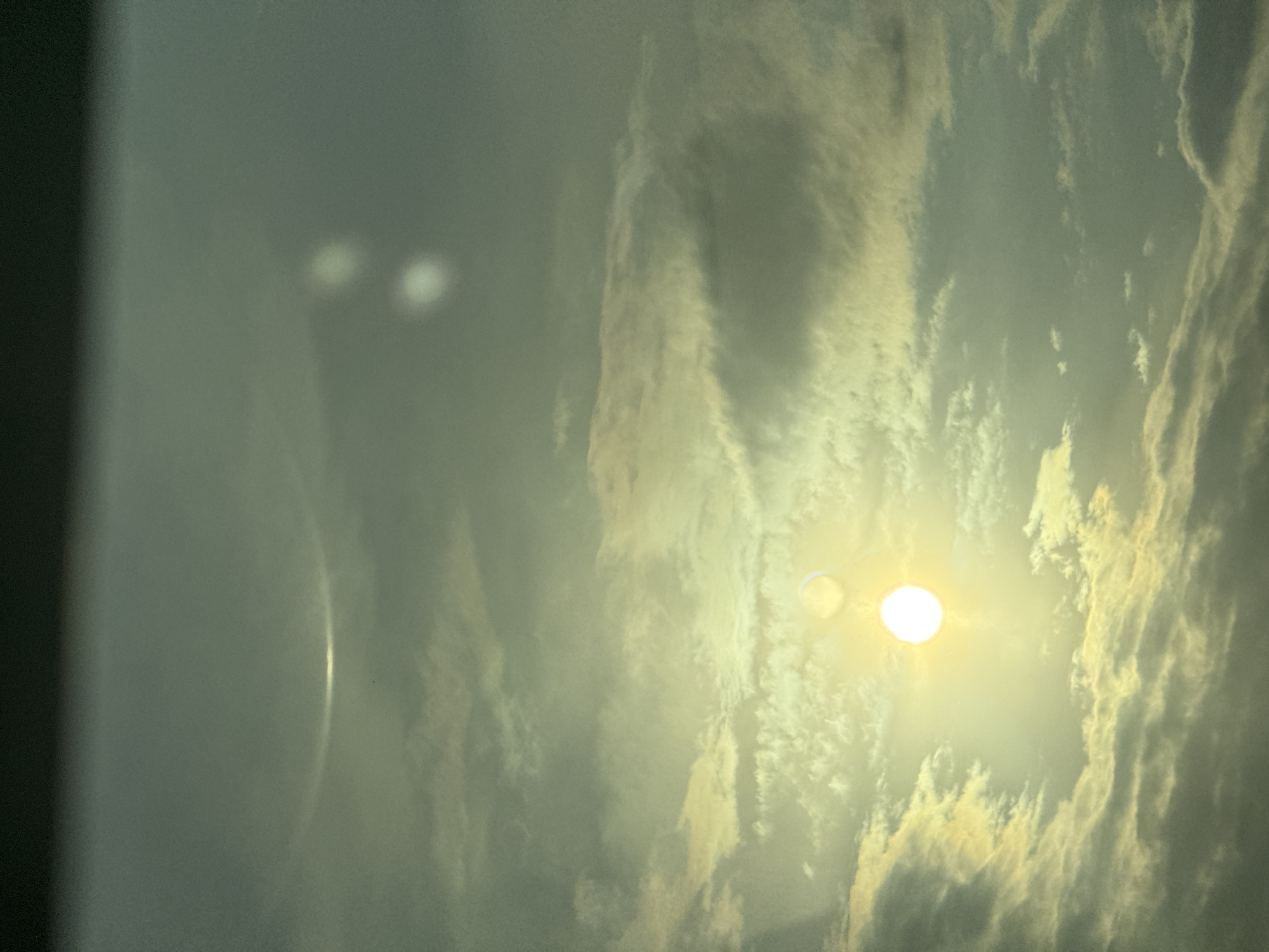 The image depicts an overcast sky observed in Suprunivka, Ukraine. A brilliant white-yellow circular sun is positioned in the lower-middle-left of the frame, emitting intense light. The sky is densely covered with stratified clouds, which are illuminated by the sun, displaying hues of green and yellow. Several atmospheric optical phenomena are visible: a prominent, luminous, curved arc extends horizontally across the upper segment of the sky; two distinct, bright, circular luminous spots, consistent with sun dogs, are located to the upper-right of the sun; and a fainter, partial arc is discernible directly above the sun. No human subjects, man-made objects, or specific actions are present in the scene.