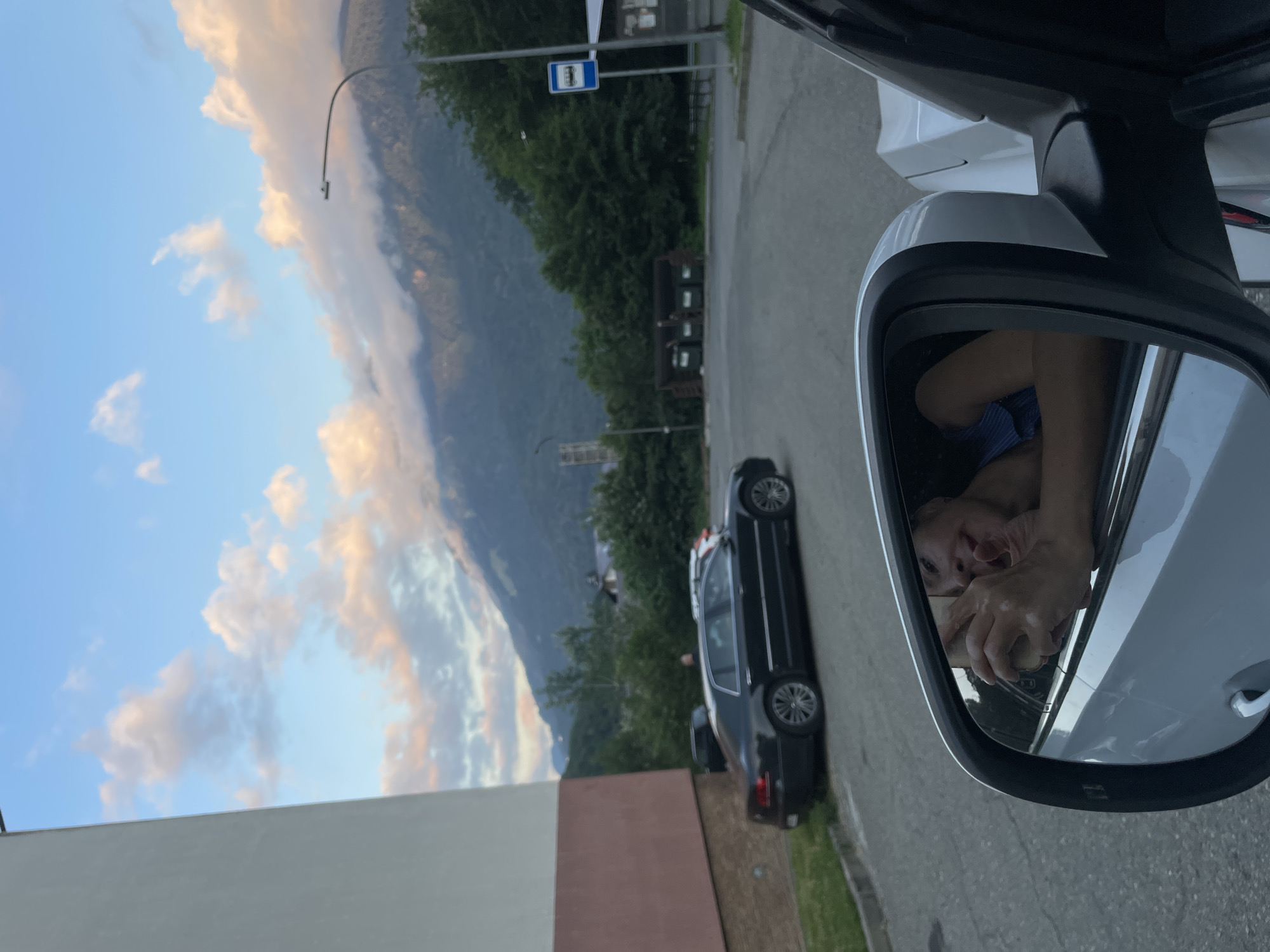 A woman is reflected in a car's side mirror, leaning out of the window. She is smiling and holding something in her hands. The reflection shows her shoulders and upper chest, and she is wearing a blue top. The car appears to be parked in an outdoor lot. In the background, mountains with clouds are visible, suggesting a location in Krasnaya Polyana, Russia. There are trees, a bus stop sign, trash receptacles, and other vehicles in the vicinity. The sky is a clear blue with some light clouds illuminated by the setting sun. A portion of a building is visible on the left side of the frame.