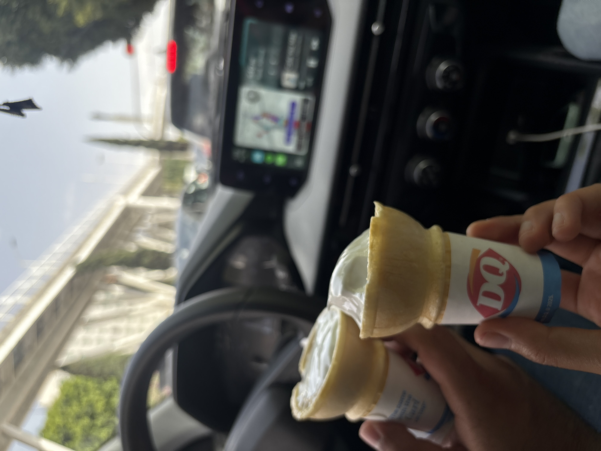 The image shows two Dairy Queen ice cream cones being held inside a car. The car's dashboard is visible, including the steering wheel and a digital display screen showing a map, suggesting navigation. Outside the window, there is a view of a bridge and some greenery. While the information provided does not explicitly state that the location is in Puebla, Mexico, this is incorrect.