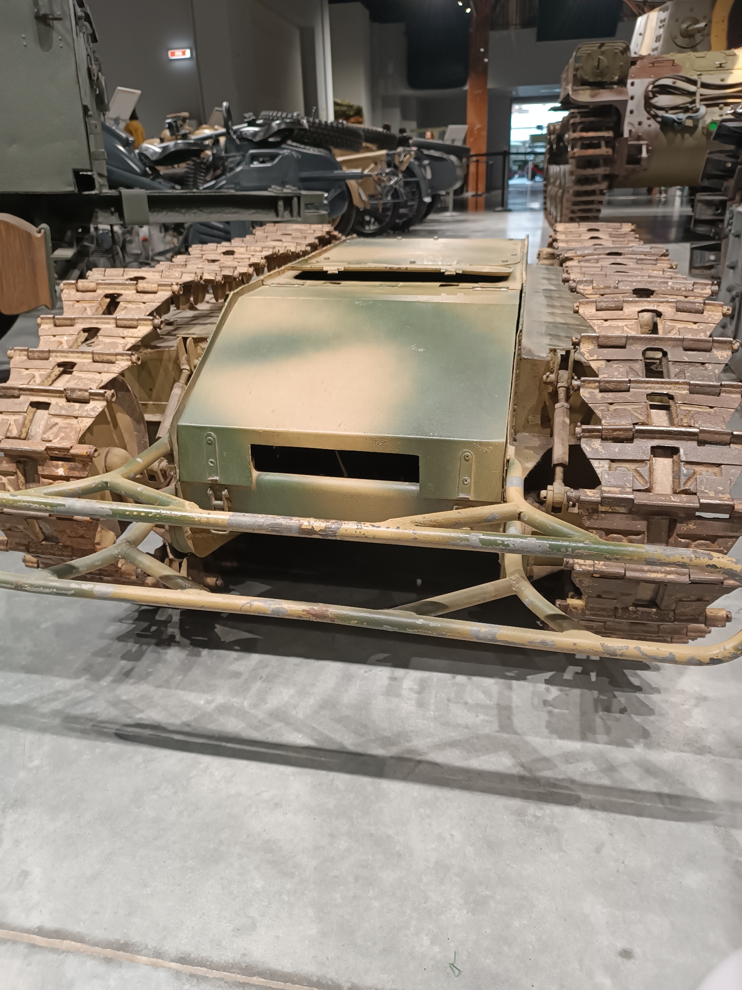 The image shows a close-up, ground-level view of a military tracked vehicle, likely a tank or armored personnel carrier, on display indoors. The vehicle is painted in a camouflage pattern of green, tan, and brown. Its tracks are visible on either side, made of interlocking metal segments. The front of the vehicle features a protective frame made of metal tubes, also painted in a weathered camouflage scheme. In the background, other military vehicles and displays can be seen, along with museum visitors. The floor is concrete, and shadows are cast from overhead lighting. The scene is consistent with an exhibit at the Military Technology Museum in Verkhnyaya Pyshma, Russia, which houses a large collection of Soviet and Russian military equipment. There are no people depicted prominently in the foreground, but their presence is implied by the museum setting.