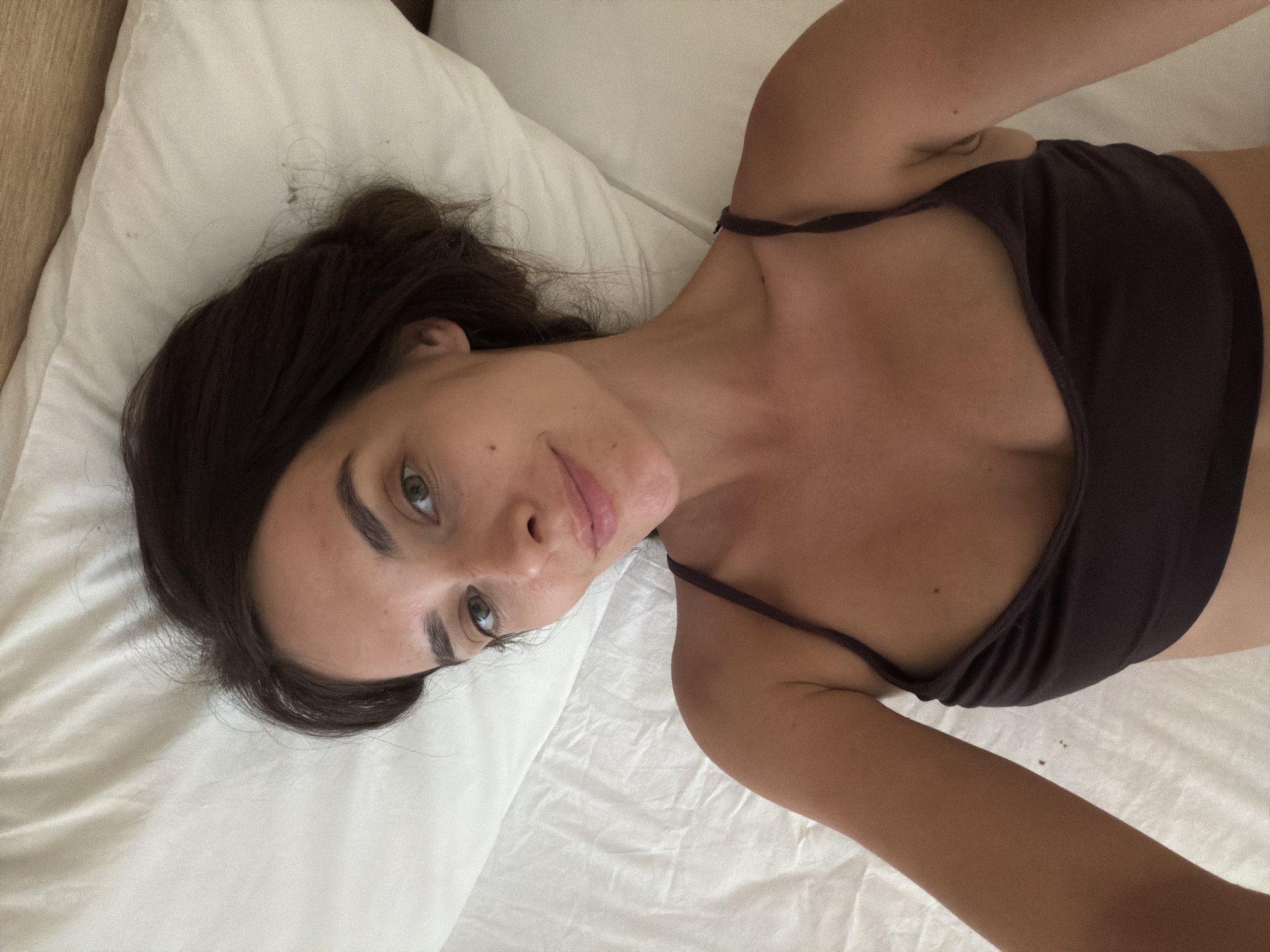 A woman with dark hair and green eyes is lying on a bed, looking at the camera. She is wearing a dark brown tank top. The bed has white sheets and a white pillow. The scene appears to be indoors, possibly in a bedroom. No specific location in Fethiye, Turkey can be determined from the image.