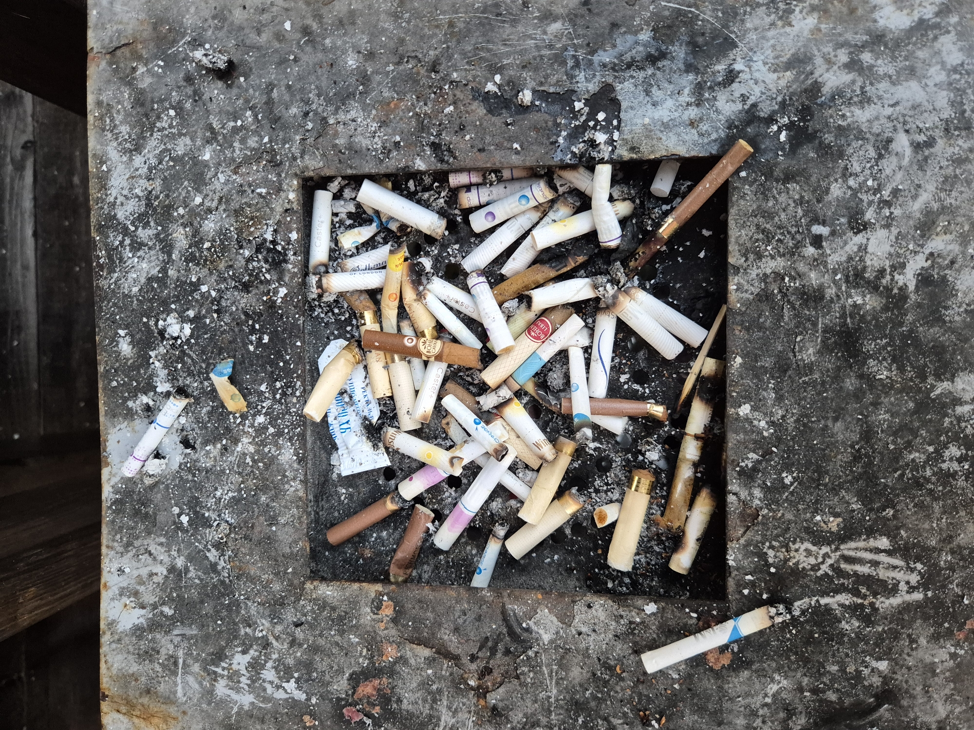 The image shows a close-up, overhead view of a square-shaped concrete ashtray filled with numerous cigarette butts. The ashtray is positioned on a weathered, dark gray surface, possibly a table or ledge, with hints of wooden planks visible at the top. The cigarette butts are of various brands and types, with some appearing to be recently smoked due to the presence of ash and smoldering ends. The surrounding concrete surface is speckled with ash and debris. The scene is devoid of any people, and no actions or interactions are depicted. The overall impression is one of litter and neglect. The provided context indicates the location is the city of Astana, Kazakhstan.