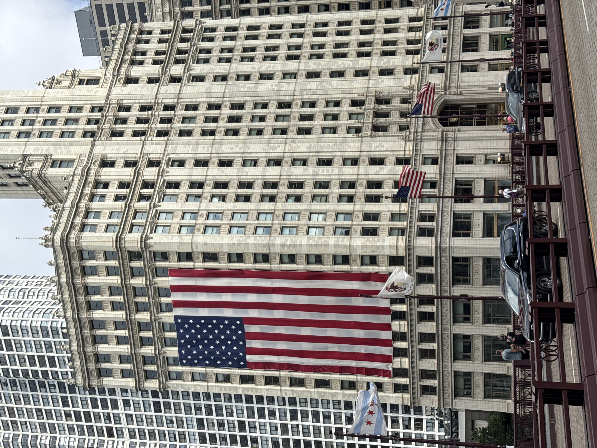 This media file shows a large, ornate building in Chicago, United States of America. The building has a facade of light-colored stone with many windows. In front of the building, an American flag is prominently displayed, hanging horizontally. To the right of the American flag, a flag with a white background and an emblem in the center is visible, possibly the flag of Illinois. Further down, a Chicago city flag with its iconic stars and stripes is also present.
The scene appears to be taking place during the daytime, under an overcast sky, suggesting potentially cooler weather. The perspective of the photo is from a slightly elevated position, looking upwards at the building. There are some people visible on balconies or platforms attached to a dark, metallic structure that runs vertically alongside the building. These individuals appear to be observing the scene below. A dark-colored car is parked on the structure. The overall impression is a view of a classic urban architectural setting, adorned with patriotic symbols.