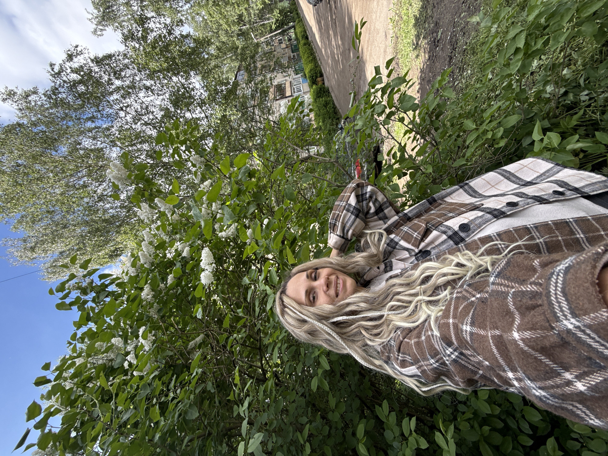 A woman with blonde hair poses in front of a lilac bush in Rusanovo, Russia. She is wearing a brown and white plaid shirt. The sky is visible in the upper left corner. A path and buildings are visible in the background.