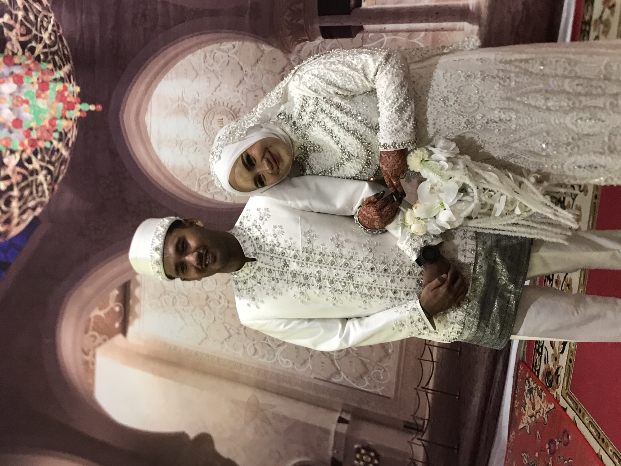 A wedding photograph features a groom and bride, likely taken in Beunyot, Indonesia. The groom, wearing a white suit with intricate silver detailing and a matching hat, stands to the right. The bride, in a matching white, heavily beaded gown and hijab, stands beside him. She holds a bouquet of white flowers. Both are smiling at the camera. The background appears to be a decorative wall with a repeating arch pattern, reminiscent of Islamic architecture, and an arrangement of colorful decorations. A red and patterned rug is visible on the lower right of the frame. The image has been rotated.