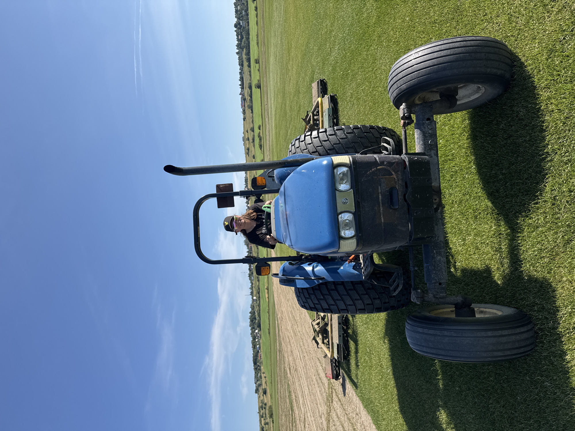 A person wearing a black t-shirt, sunglasses, and a black baseball cap is operating a blue tractor on a bright, sunny day. The tractor is equipped with mowing attachments on the rear. The scene appears to be a large, well-maintained grassy area, possibly a field or a park, with a section of cut grass visible in the background. In the distance, there are rolling hills with scattered houses and trees under a clear blue sky with a few wispy clouds. The image captures the tractor from a low-angle, frontal view, emphasizing its large tires and the person driving it.