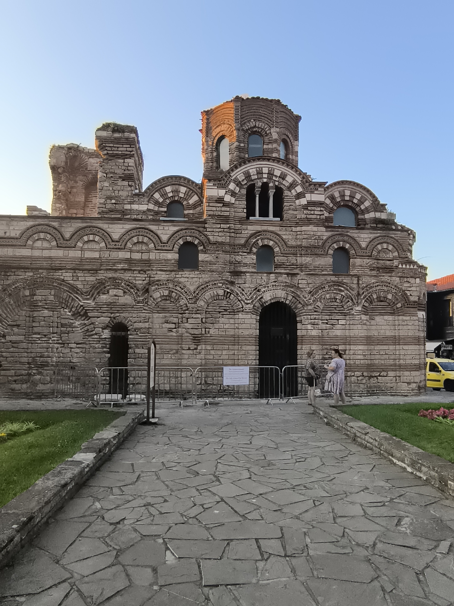 The image shows the Church of St. Stephen in Nesebar, Bulgaria, viewed from a courtyard paved with irregular stone slabs. The church is a large, ancient stone structure with multiple arched windows and decorative brickwork. The facade is weathered and shows signs of age, with some sections appearing to be in ruins. Two women stand near the entrance, one in a patterned shirt and dark skirt, the other in a light-colored dress. A metal barrier is set up in front of the church entrance. Beyond the church, a yellow car is visible parked on a street. Green grass borders the courtyard, and some pink flowers are visible on the right side of the image. The sky is clear and blue, indicating it is daytime.