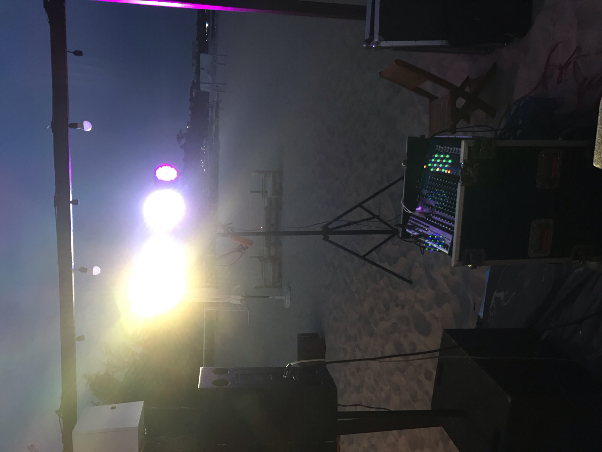 The image depicts an outdoor event setup on a sandy beach at dusk. The scene is dominated by artificial lighting, with a bright, intense light source on the left creating a hazy effect, possibly due to fog or smoke. There are several lights visible, including a prominent bright white light and a purple light. In the foreground, a DJ mixing console is set up on a case, with its illuminated buttons suggesting active use. Speakers and audio equipment are also present, indicating a musical performance or event. A wooden chair is positioned behind the DJ setup, suggesting a waiting area or a place for the performer to sit. The background reveals a hazy view of structures and lights, hinting at a populated area or beachfront facilities. The sand-covered ground and the style of lighting suggest a beach party or an outdoor concert. The time of day is evident from the dark sky, indicating evening or late afternoon. There are no visible people in the frame. The overall impression is that of a vibrant, yet tranquil, evening gathering being set up or in progress.