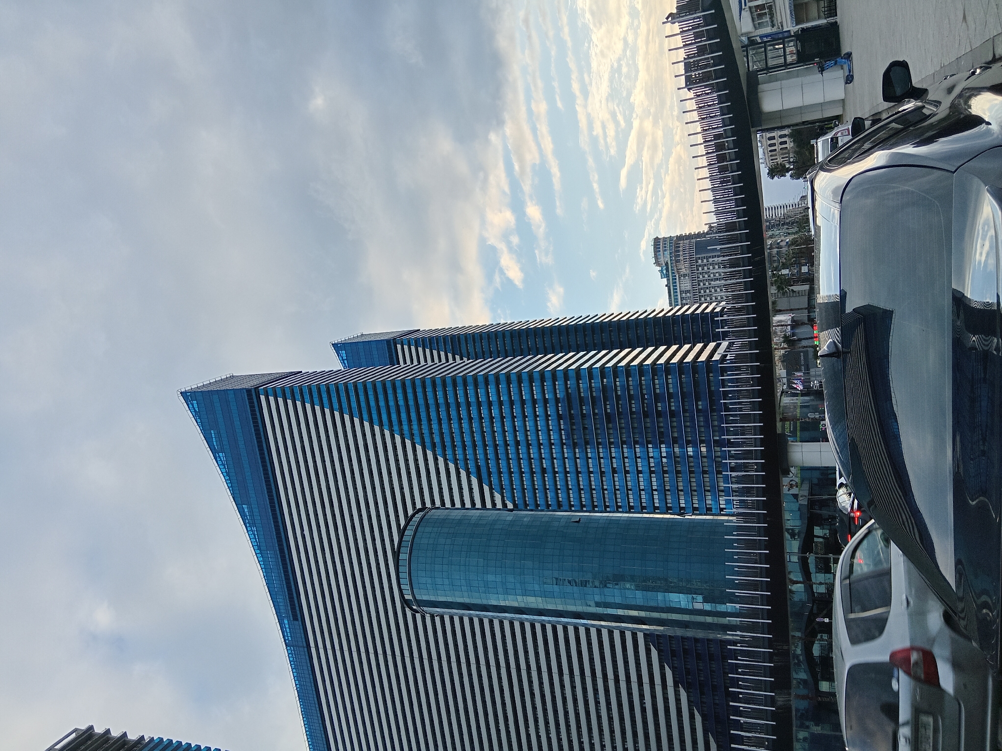 The photo depicts the Radisson Blu Hotel, a striking skyscraper in Batumi, Georgia. The hotel features a modern design with blue-tinted glass and white horizontal stripes. A cylindrical glass structure protrudes from the side of one of the building segments. Cars, including a dark-colored SUV, are visible in the foreground, suggesting a street scene near the hotel. The sky is partly cloudy with visible buildings in the distance.