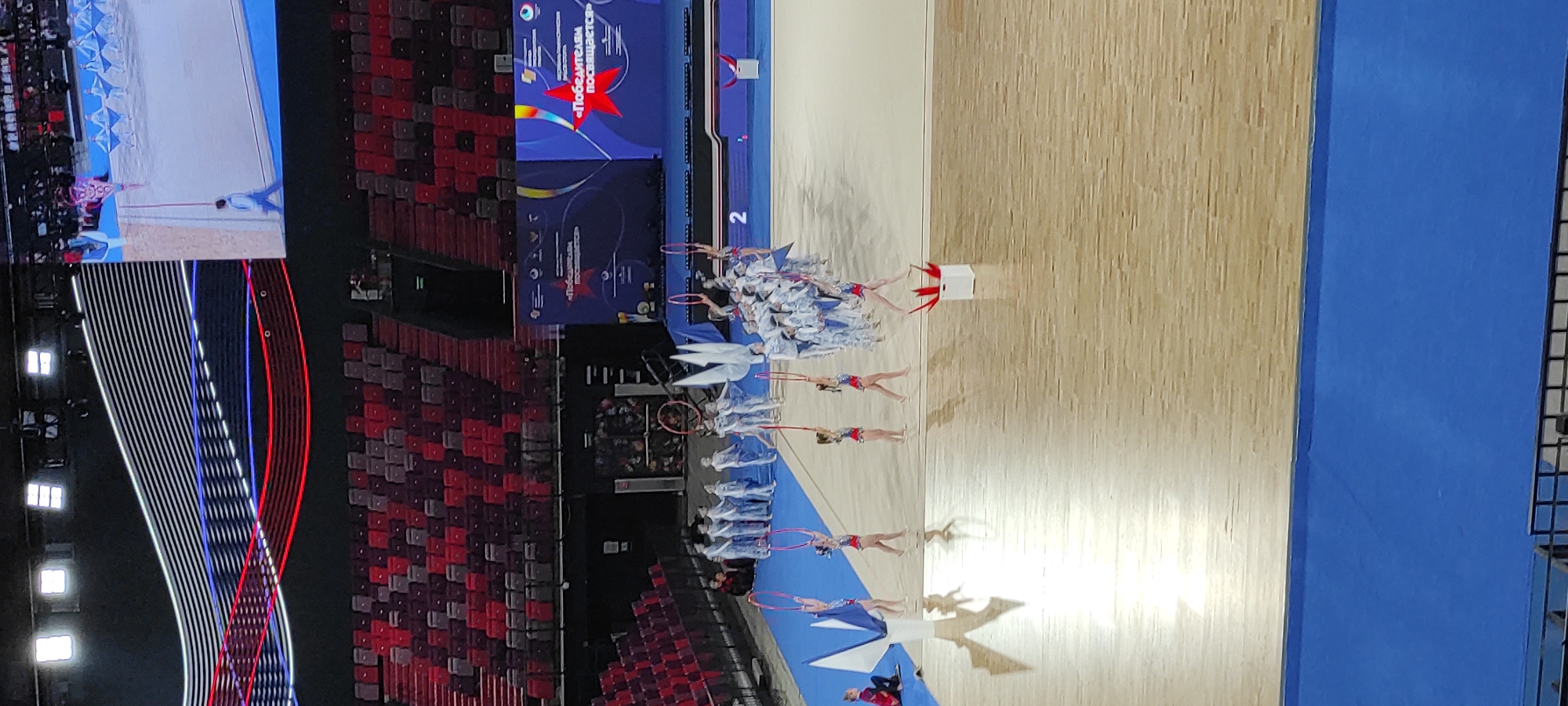 The image depicts a rhythmic gymnastics competition in Moscow, Russia. The scene is set in a large indoor arena with tiered red seating in the background. A large screen displays graphics and text, including a prominent red star and Russian lettering. On the blue and beige gymnasium floor, a group of young rhythmic gymnasts in matching white and blue outfits are performing with hoops and flags. Some gymnasts are in mid-performance, while others stand in formation. The lighting is bright, typical of an indoor sporting event. The overall atmosphere suggests a formal competition or performance.