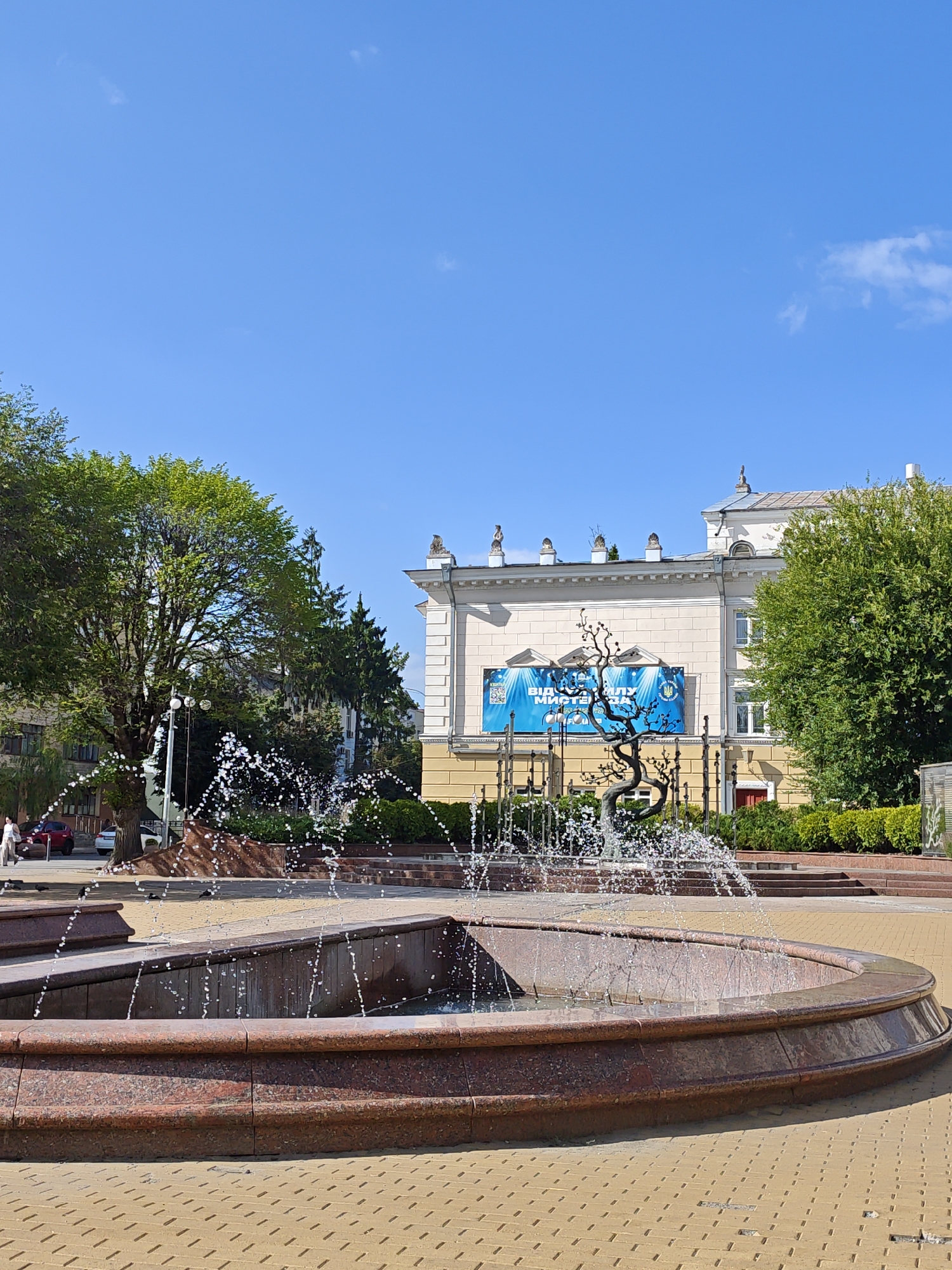 The image depicts a public square with a fountain in the foreground. The fountain has a multi-tiered, granite basin with water actively spraying upwards, creating a misty effect. Behind the fountain, a bronze sculpture of a stylized tree stands. Further back, a light-colored building with a prominent blue banner displaying Cyrillic text and a Ukrainian trident emblem is visible. Trees and green landscaping surround the building and the square. The ground is paved with yellow interlocking bricks. In the background, several cars and two individuals are present on a street. The sky is clear and blue with a few wispy clouds. This scene is located in Vinnytsia, Ukraine, at the Theater Square, specifically in front of the Vinnytsia Regional Ukrainian Academic Music and Drama Theater.
