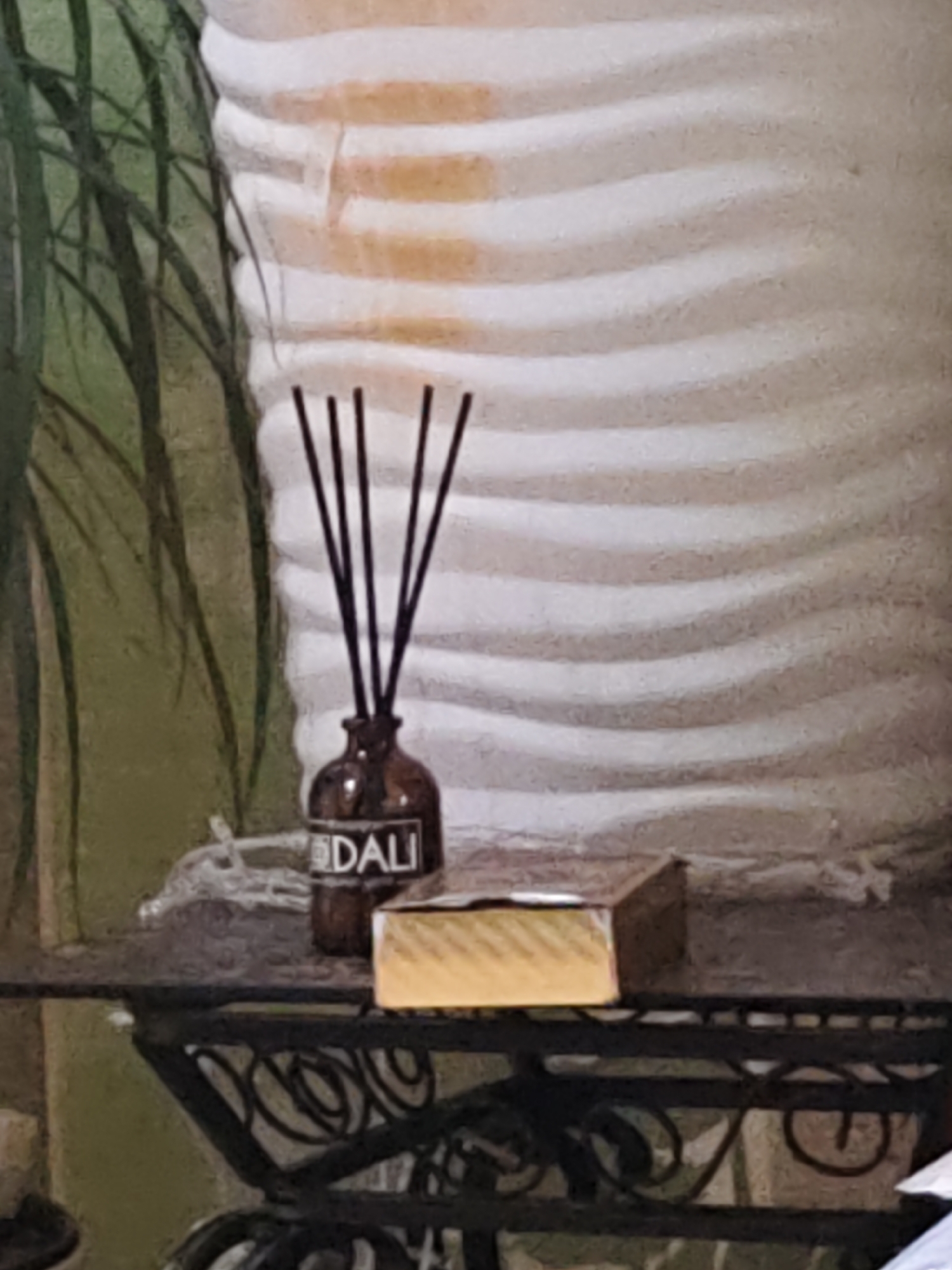 This is a still life image featuring a diffuser with reeds in a dark amber bottle labeled "DALI" and a rectangular gold box on a dark metal shelf. The background consists of a large, ribbed off-white structure and some out-of-focus green foliage. The lighting appears to be natural, casting soft shadows. The location in Olginskaya, Russia is not discernible from the image itself.