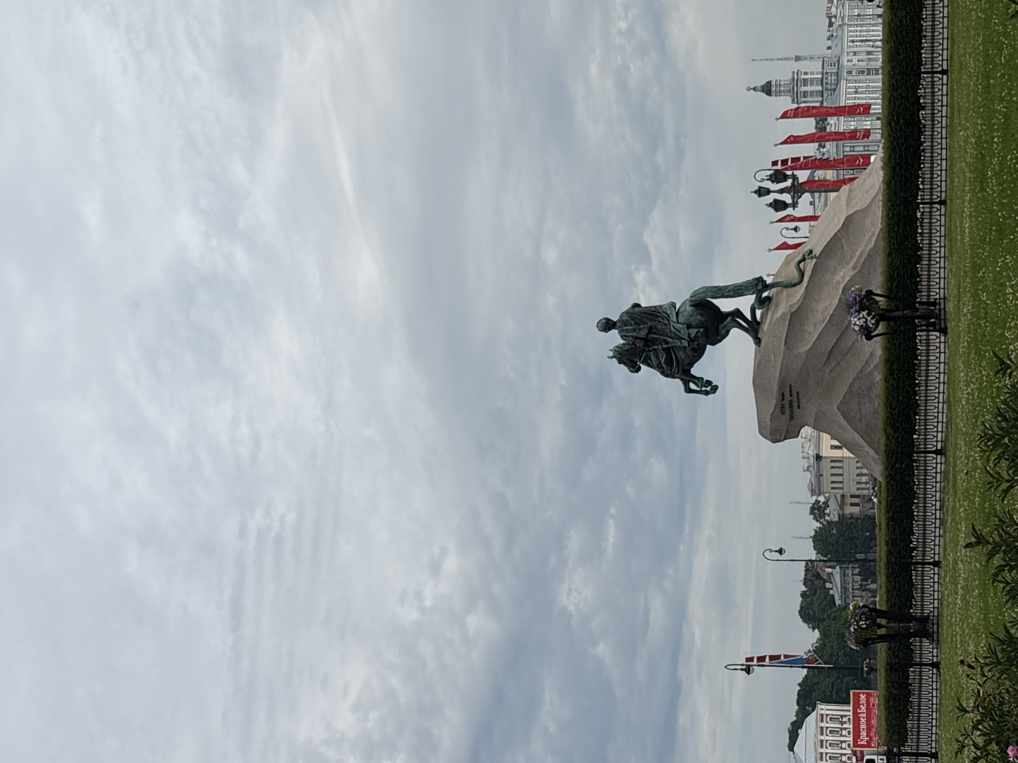 The image captures the Bronze Horseman, a monument to Peter the Great, located in Senate Square in Saint Petersburg, Russia. The equestrian statue, crafted from bronze, stands dramatically atop a massive granite boulder, known as the Thunder Stone. The horse is depicted rearing on its hind legs, while Peter the Great, in motion, looks onward. A snake is shown beneath the horse's hooves, symbolizing the victory over his enemies.
In the background, characteristic architecture of Saint Petersburg is visible, including classical buildings with domes and ornate facades. Red flags with white lettering are prominently displayed, adding a splash of color against the overcast sky. The sky itself is filled with a layer of textured, gray clouds, suggesting a temperate or possibly cooler day.
In the foreground, a manicured lawn with scattered white flowers leads up to a black metal fence. Beyond the fence, there are hedges and decorative planters filled with flowers. A street lamp with multiple lights stands near the monument. To the left, a large red banner with white text that reads "Красное&Белое" (Krasnoe & Beloe, a Russian liquor store chain) is visible, indicating a commercial presence in the vicinity. The overall scene is one of historical significance and urban landscape, characterized by grand architecture and a notable monument.