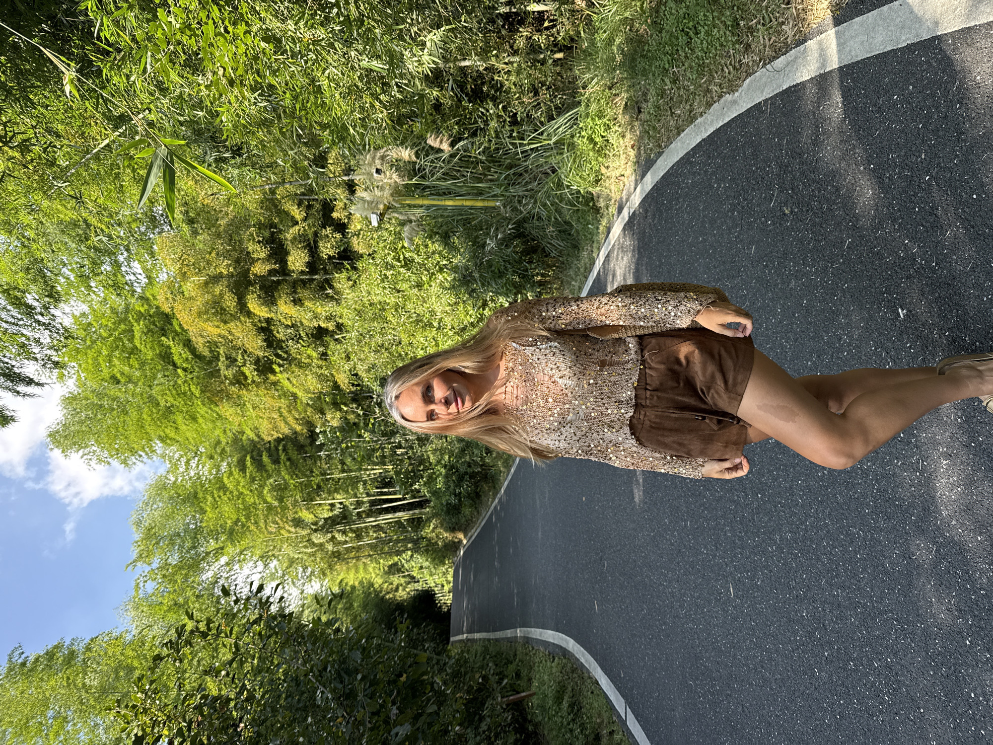 A blonde woman stands on a paved, curving road bordered by dense green foliage, likely bamboo, under a bright sky with scattered clouds. The woman is positioned in the foreground, facing slightly towards the camera. She is wearing a sequined, long-sleeved top and brown shorts. A bag is slung over her shoulder. The road appears to be a pathway or scenic route within a natural environment, possibly a park or botanical garden in Wuxing District, China, given the dense bamboo growth. The sun casts shadows on the road, indicating it is daytime.