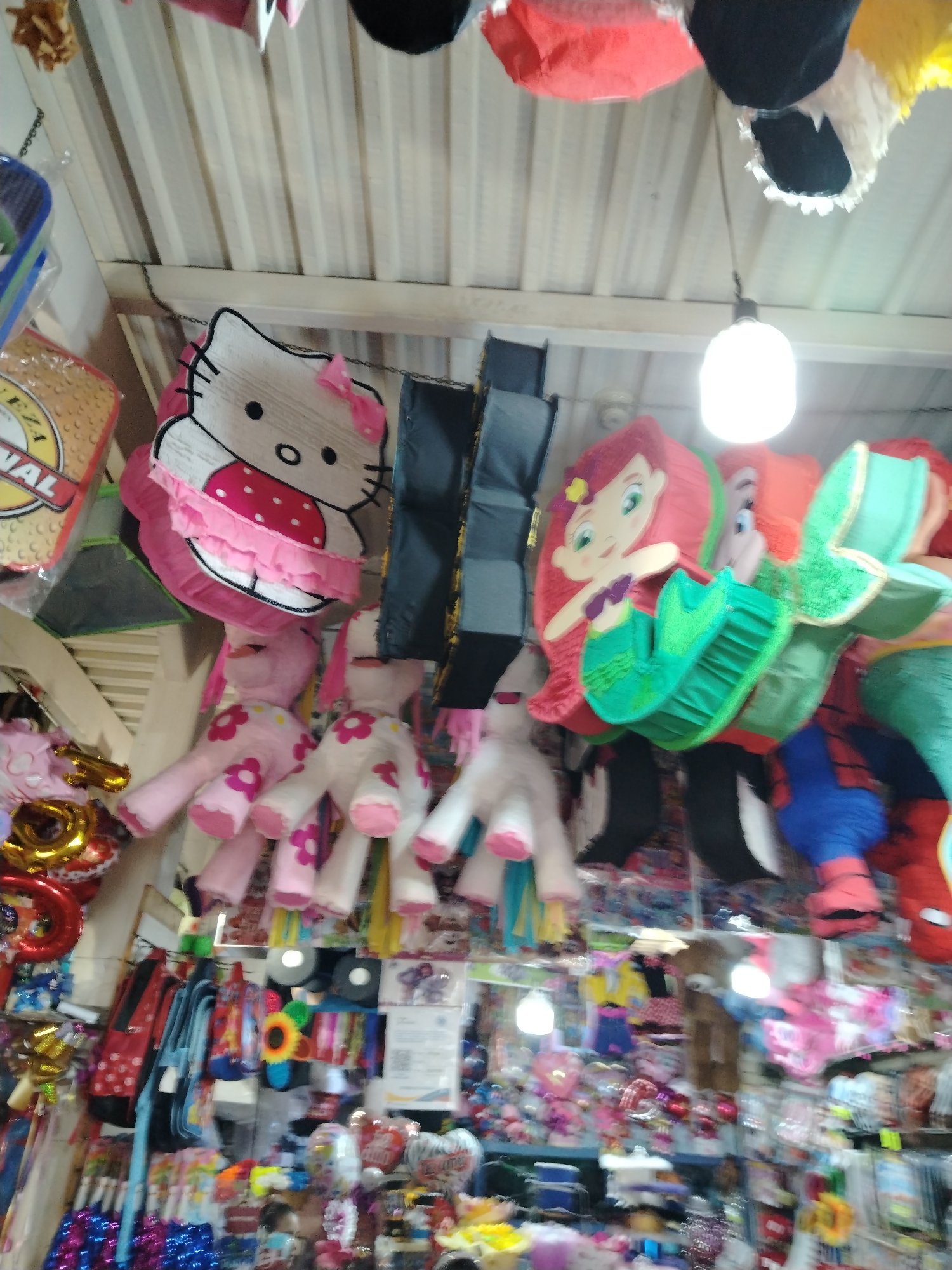 The image depicts the interior of a retail shop in Tinaquillo, Venezuela. A variety of merchandise hangs from the ceiling, including character-themed piñatas (Hello Kitty, Ariel from The Little Mermaid, and Spiderman) and plush toys. Below, shelves and racks are stocked with items such as backpacks, toys, balloons, and various accessories. The ceiling is made of corrugated metal, and a light fixture illuminates the scene.