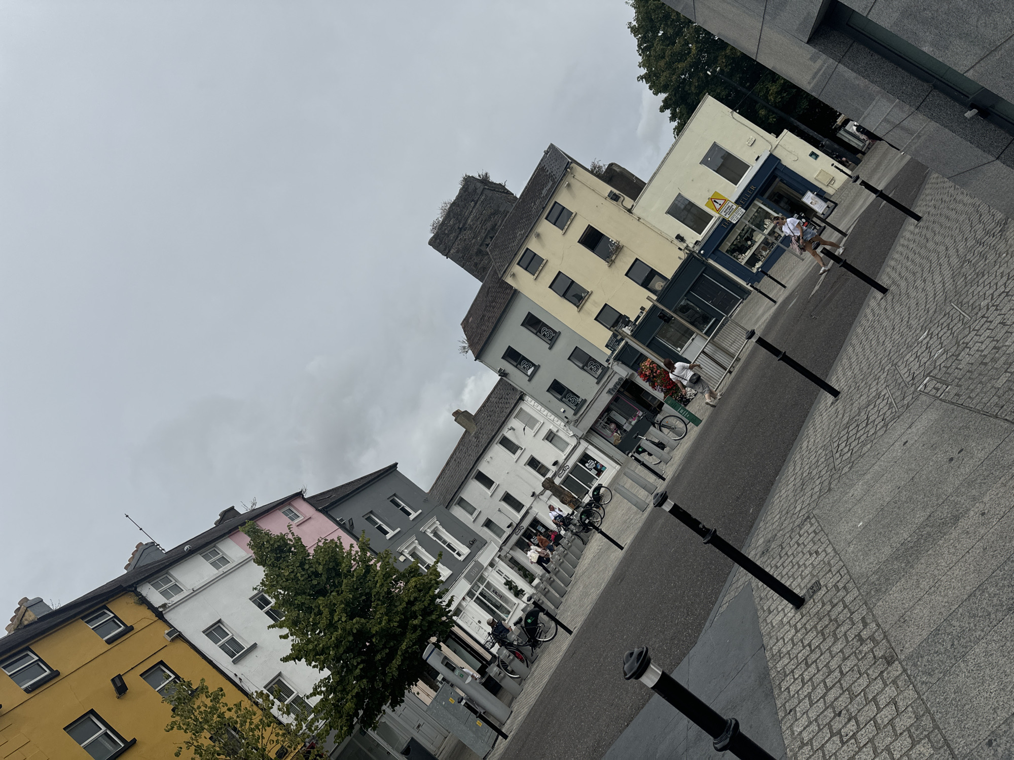 The image depicts a street scene in Waterford, Ireland, with buildings lining one side of the road. The buildings are of varying heights and colors, including yellow, white, pink, and gray facades. Some of the buildings appear to be shops, with visible storefronts and signage. There are people walking and cycling on the street and sidewalk. Several bicycles are parked along the street. The sky is overcast. The foreground shows a cobblestone sidewalk and a paved road. Bollards are placed along the sidewalk. A tree is visible near the buildings. The image is taken at an angle, with the buildings appearing tilted.