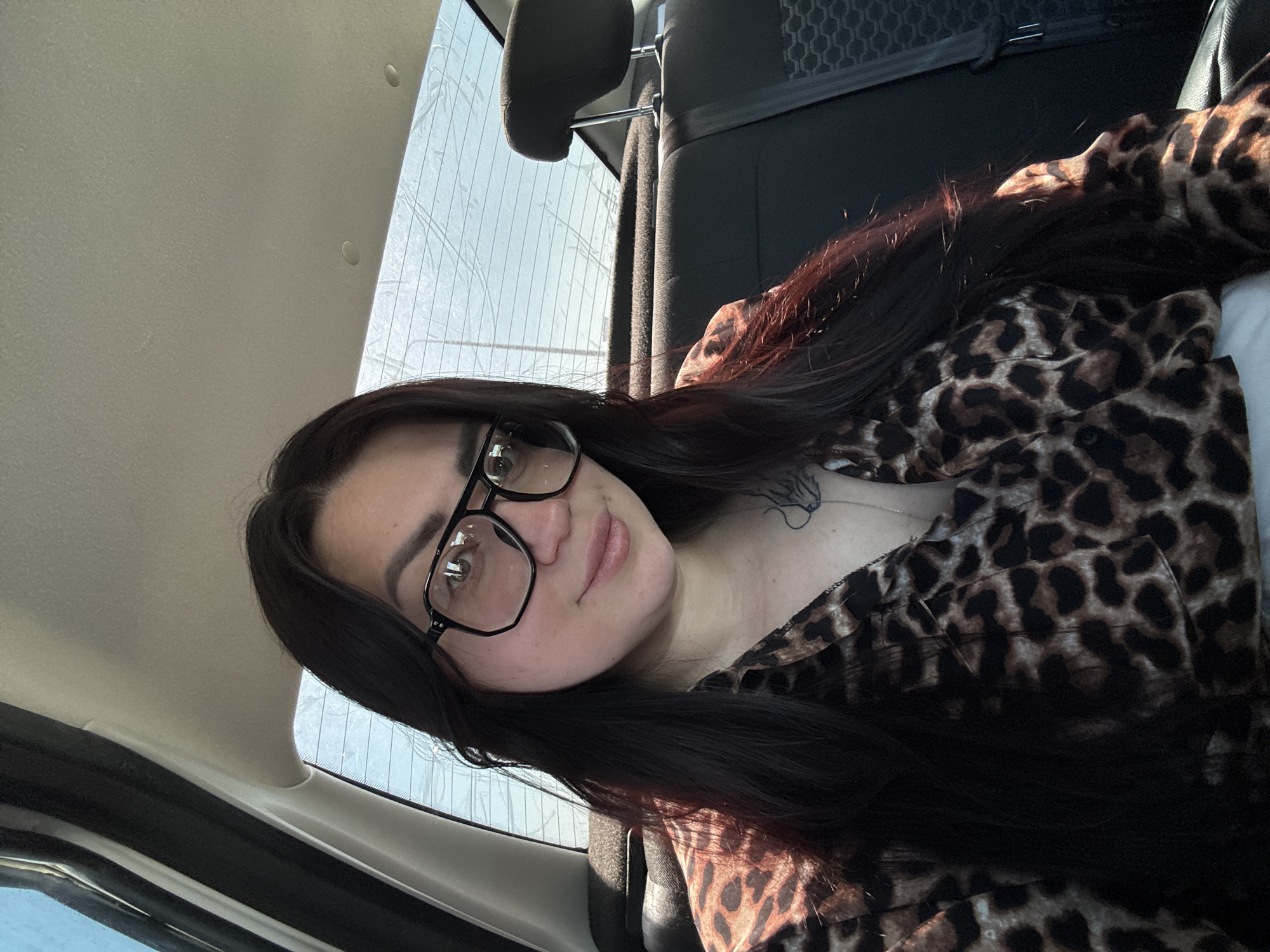 A selfie shows a woman with dark hair and glasses, wearing a leopard-print shirt, inside a car. The location is Unknown, Unknown. The headrest of the car seat is visible behind her. The car window shows an undefined outdoor scene. The woman is looking at the camera with a neutral expression.