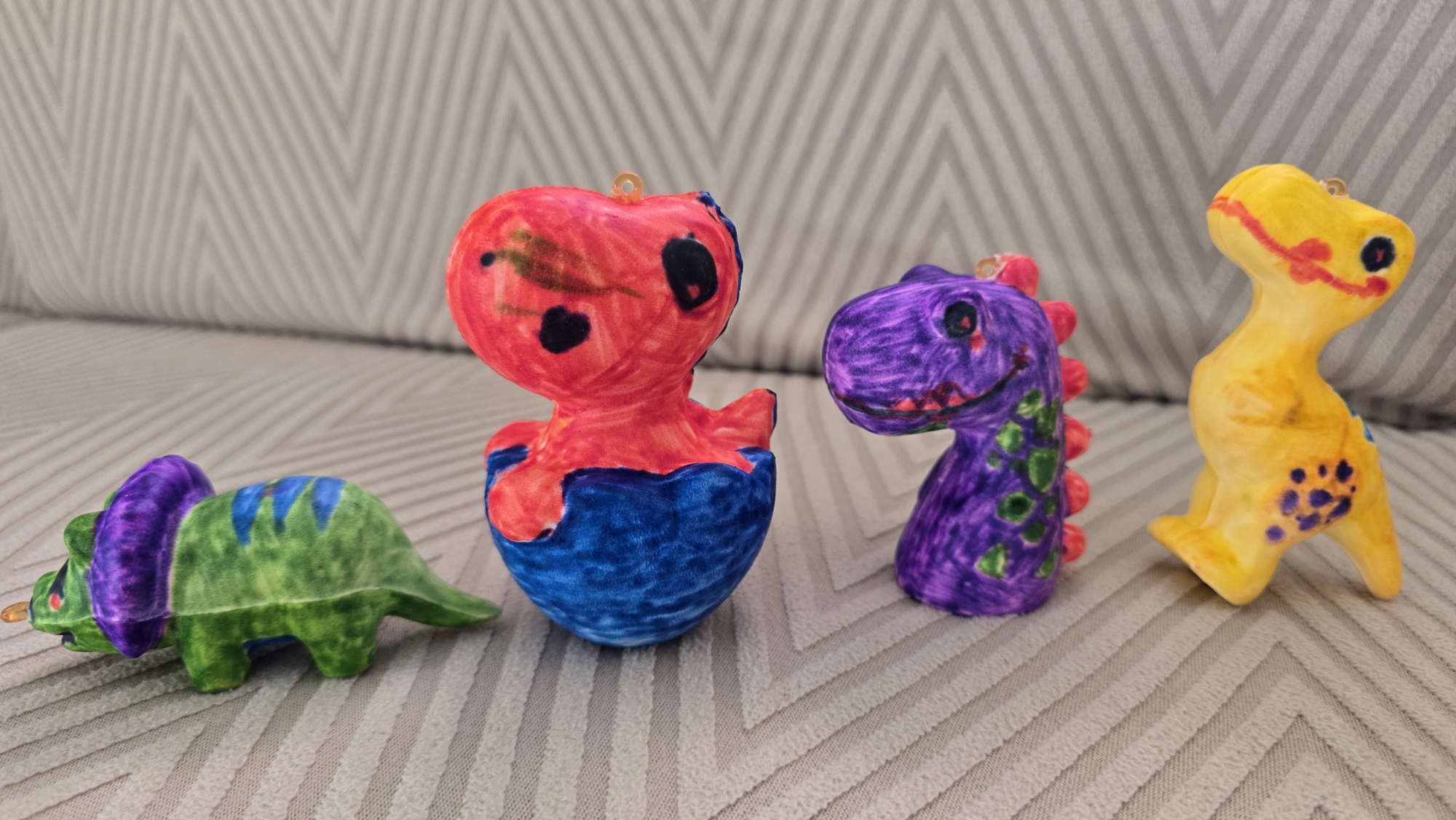 Four colorful, hand-painted ceramic dinosaur figurines are arranged on a textured gray couch in a horizontal line. From left to right, the first figurine is a green triceratops with a purple frill and blue stripes on its back. The second is a red and blue dinosaur emerging from an eggshell, with black dots for eyes. The third is a purple dinosaur with pink spikes and green spots. The fourth is a yellow dinosaur with black spots and red accents. The figurines appear to be children's crafts. The background is a gray couch with a chevron pattern. There is no indication of the location being in Верхнеказымский, Russia.