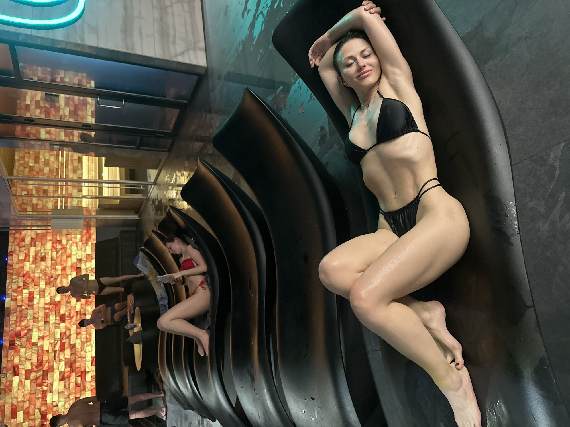 The image shows a woman in a black bikini reclining on a curved, black chaise lounge. Her arms are raised above her head, and her eyes are closed. She appears relaxed. The setting appears to be a spa or wellness center, as several other people are seen in the background also lounging on similar chairs. The background features a wall of salt bricks and a teal-colored light fixture. Based on the spa's design, it is likely taken at the Grand Fontana Hotel in Saint Petersburg, Russia.