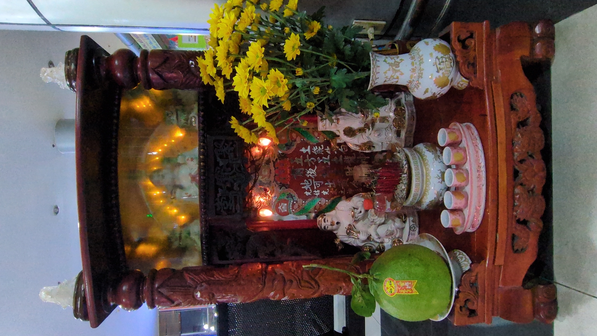 A tilted, close-up shot depicts a domestic altar in Da Lat, Vietnam. The altar, made of dark wood with ornate carvings, is adorned with various religious and symbolic objects. On the altar are statues, vases of yellow flowers, and a set of small cups. A large, green pomelo sits on a plate at the front of the altar. In the background, there is a reflective surface displaying small lights. The altar is set against a backdrop of interior elements, including a wall.