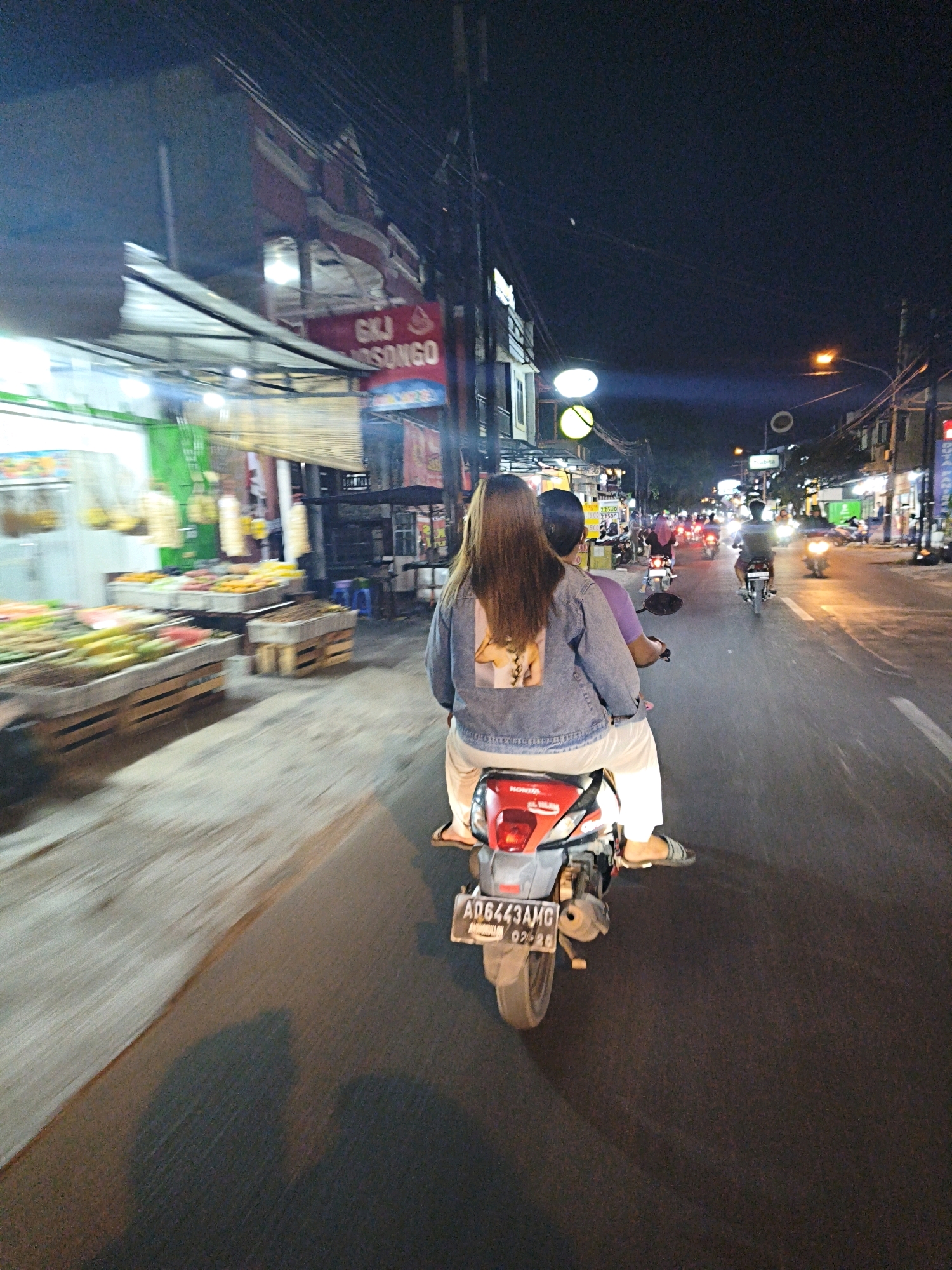 In the city of Surakarta, Indonesia, at night, two individuals are riding a motorcycle. The person in the back is wearing a denim jacket with a graphic print on the back and white pants. The person in front is wearing a purple shirt. They are traveling on a street lined with shops and market stalls. The market stalls on the left display an assortment of fruits and vegetables. The street is illuminated by streetlights and the lights from the shops and passing vehicles. Several other motorcycles are visible on the road ahead, also in motion. The overall atmosphere suggests a lively street scene at night in a bustling area.